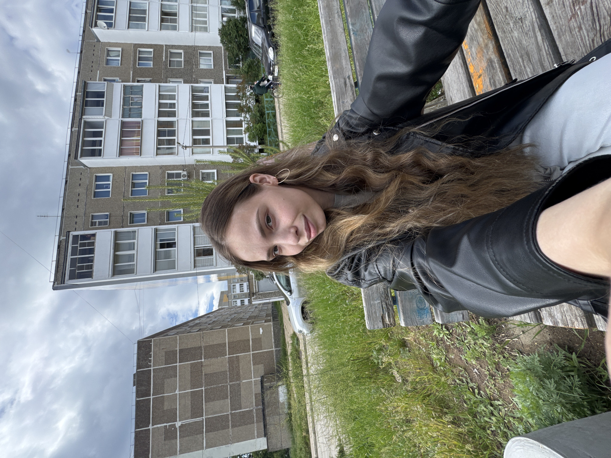 A young woman with long, light brown wavy hair and wearing a black leather jacket is sitting on a wooden bench outdoors. She is looking at the camera with a slight smile. The background features an urban landscape, including multi-story apartment buildings with numerous windows. There is also green grass and some greenery visible. A grey and white car is parked nearby. Based on the style of the buildings, this photo appears to be taken in a residential area in the city of Chaikovsky, Russia.