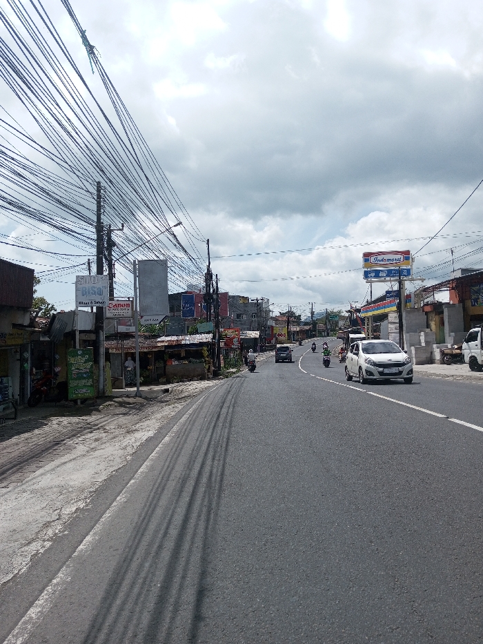 The image depicts an urban street scene in Gubbisamamidi, India, under an overcast sky. The multi-lane asphalt road features a solid white line on its left edge and a dashed white centerline. Shadows from overhead wires are cast prominently across the left lane.
**Traffic and Objects:** A white compact car travels in the right lane, heading away from the viewer. Behind it, a dark-colored sedan follows. Multiple motorcycles are present on the road, with at least four visible, interspersed among the cars and moving in various directions, primarily away from the viewer. A portion of a white utility truck is visible on the far right.
**Left Roadside:** The left side of the road features a concrete sidewalk bordered by a row of single-story commercial structures, some with corrugated metal roofs. Prominent signage includes "SHOP Bisa" and a "Canon" logo. A dense network of black utility wires is strung between numerous dark utility poles along this side of the street. Two individuals are visible near the shops; one person in light-colored clothing and a head covering stands near an entrance.
**Right Roadside:** On the right, a large red, blue, and yellow "Indomaret" sign is prominently displayed, indicating a convenience store, a chain typically found in Indonesia. Behind this sign, a petrol station canopy is partially visible. Other commercial buildings line this side of the street. Green foliage is visible in the background and behind some structures.