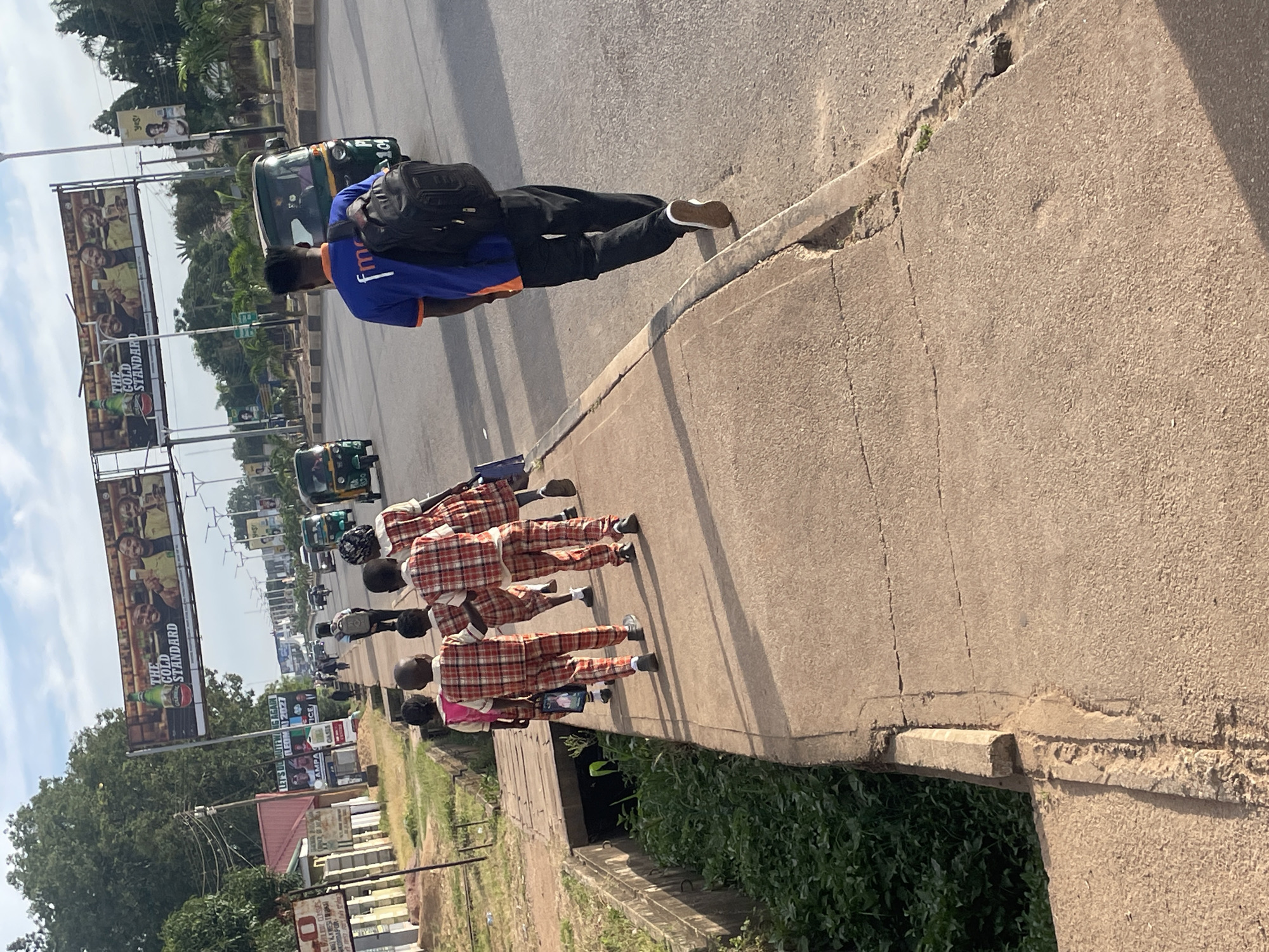 In the city of Jos, Nigeria, several schoolchildren in matching plaid uniforms are walking along a cracked concrete sidewalk. A young man wearing a blue shirt with "fmc" logo and black pants with a backpack slung over his shoulder is walking away from the camera on the adjacent road. Various vehicles, including auto-rickshaws and cars, are visible on the road, and large billboards advertising "THE GOLD STANDARD" are displayed overhead. The environment appears to be an urban setting with trees and buildings lining the street.