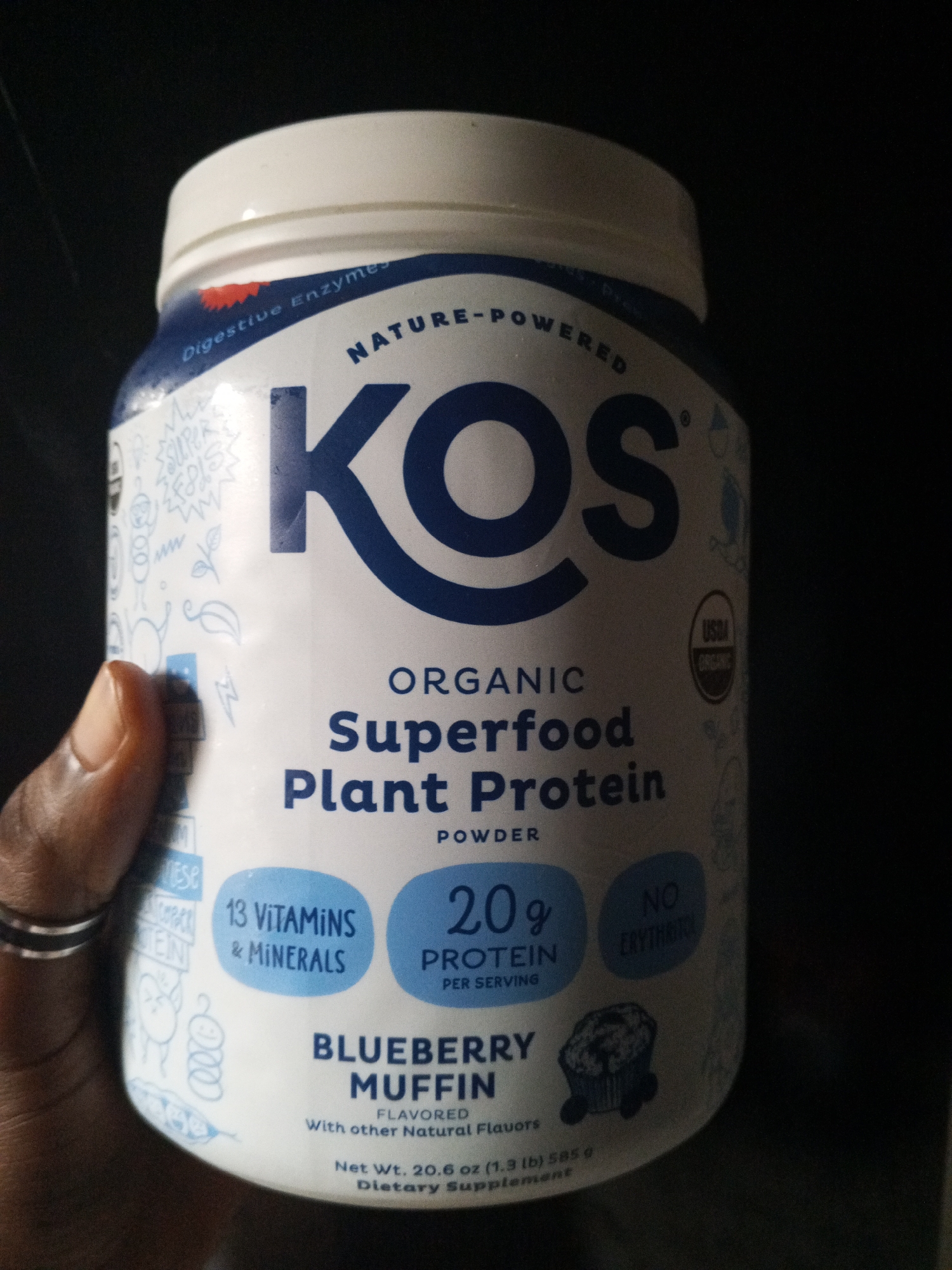 The image shows a hand holding a white and blue plastic container of KOS Organic Superfood Plant Protein powder, flavored Blueberry Muffin. The container has text and graphics indicating it is nature-powered, contains digestive enzymes, 13 vitamins & minerals, 20g protein per serving, and no erythritol. The container also has a USDA Organic seal. The background is dark and appears to be an indoor setting. The image does not provide any information about the location in the city of Ashata, Nigeria.