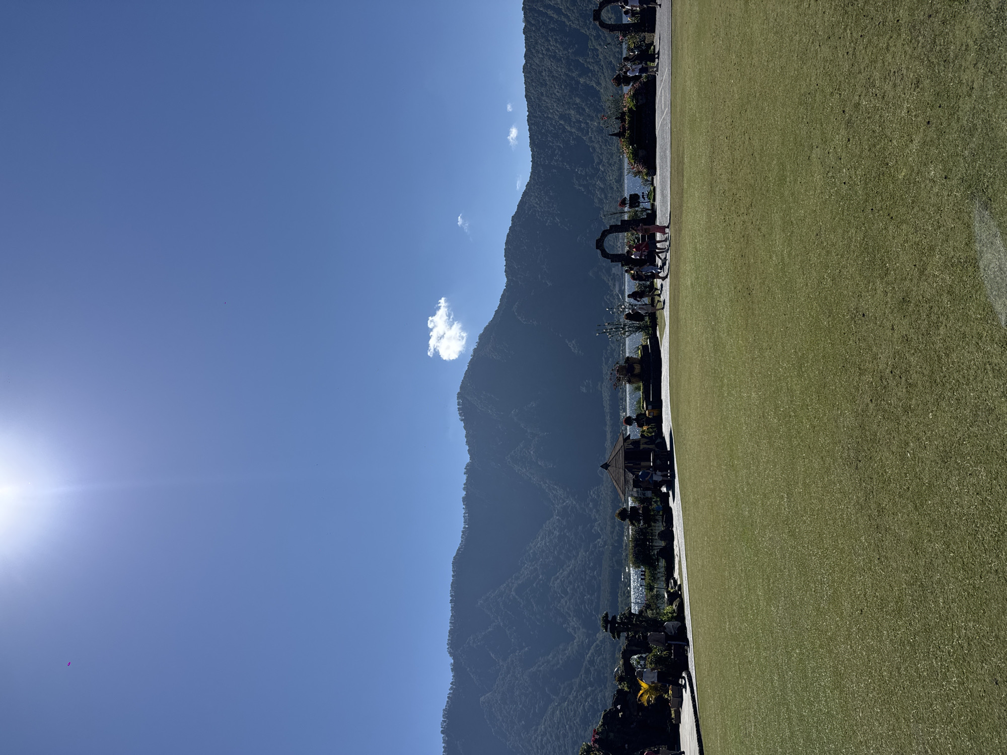 The image depicts a scenic landscape with a large, well-maintained grassy field in the foreground. In the middle ground, a paved area with several people walking is visible, leading to a body of water. Beyond the water, lush green mountains rise majestically, their slopes covered in dense vegetation. The sky is a vibrant blue, with a few scattered white clouds. A bright sun is positioned in the upper left corner, casting a warm glow over the scene.
The location is identified as Baturiti, Indonesia. The scene appears to be a park or a tourist attraction, given the presence of manicured lawns, decorative archways, and people engaged in leisurely activities. The overall atmosphere is serene and picturesque, highlighting the natural beauty of the area and the peaceful ambiance of the day.
