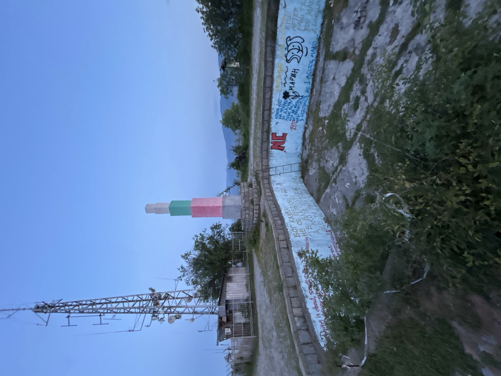 The image shows the Monument of Bulgarian-Soviet Friendship, a controversial monument located on Bunardzhik Hill in Plovdiv, Bulgaria. The monument is a tall, multi-colored structure with a tower composed of green, white, and pink blocks. An adjacent low concrete wall is covered with graffiti, including phrases written in Bulgarian and English, as well as illustrations. A telecommunications tower stands to the left of the monument. The sky is a light blue, suggesting either early morning or late evening. The environment is a mix of built structures, trees, and shrubs, which are a part of the natural landscape.