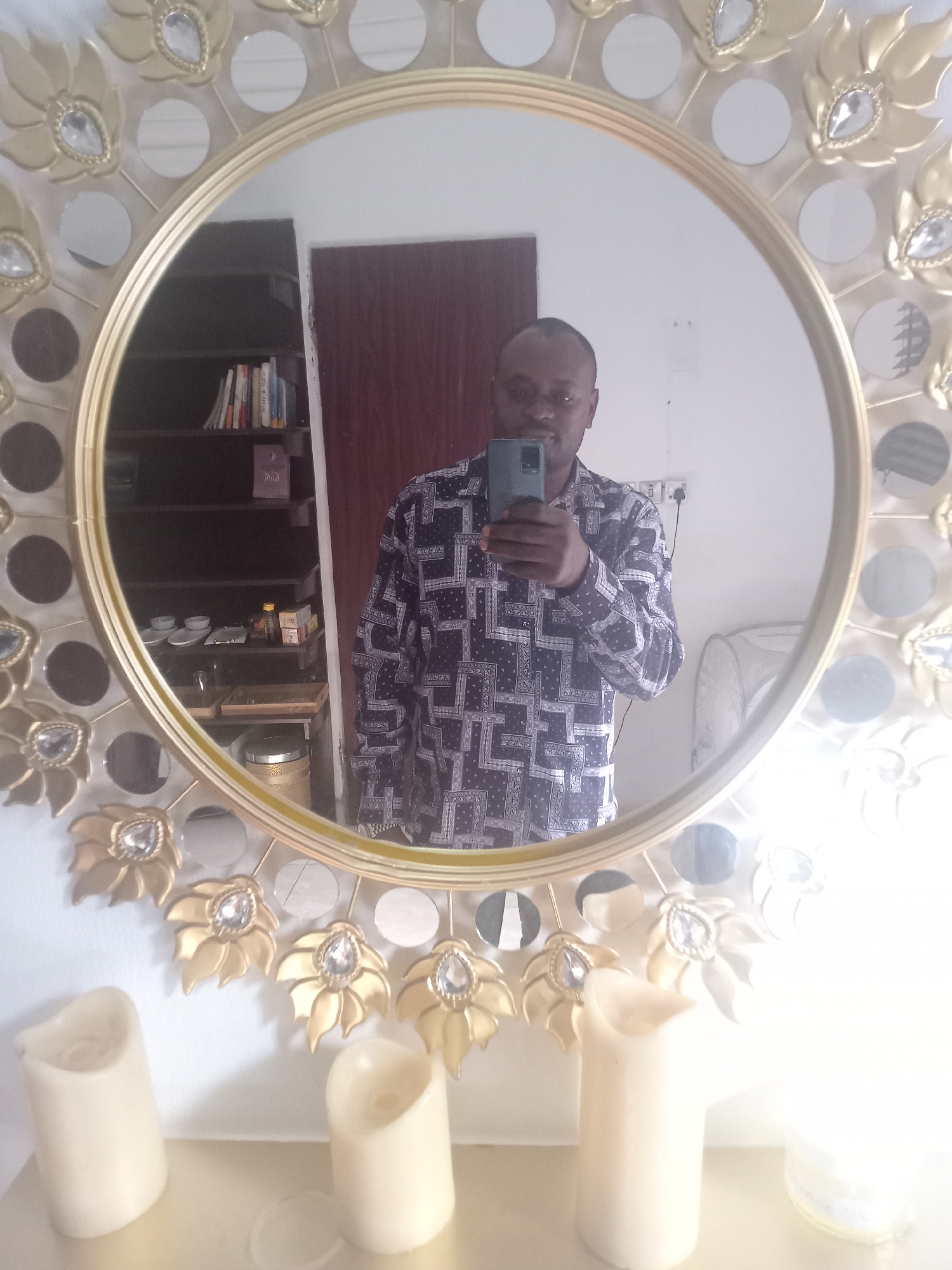 A man is taking a selfie in a decorative, round mirror in what appears to be a room in a building in Gwarinpa, Nigeria. The mirror has a gold frame adorned with decorative gold leaves and clear gem-like embellishments. Reflected in the mirror is the man holding a smartphone, a dark brown door, a bookshelf containing books and various objects, and part of a light-colored armchair or sofa. In front of the mirror, on what appears to be a shelf, are four cylindrical, off-white candles. The room's walls are white, and there are electrical outlets visible near the top of the wall in the mirror's reflection.
