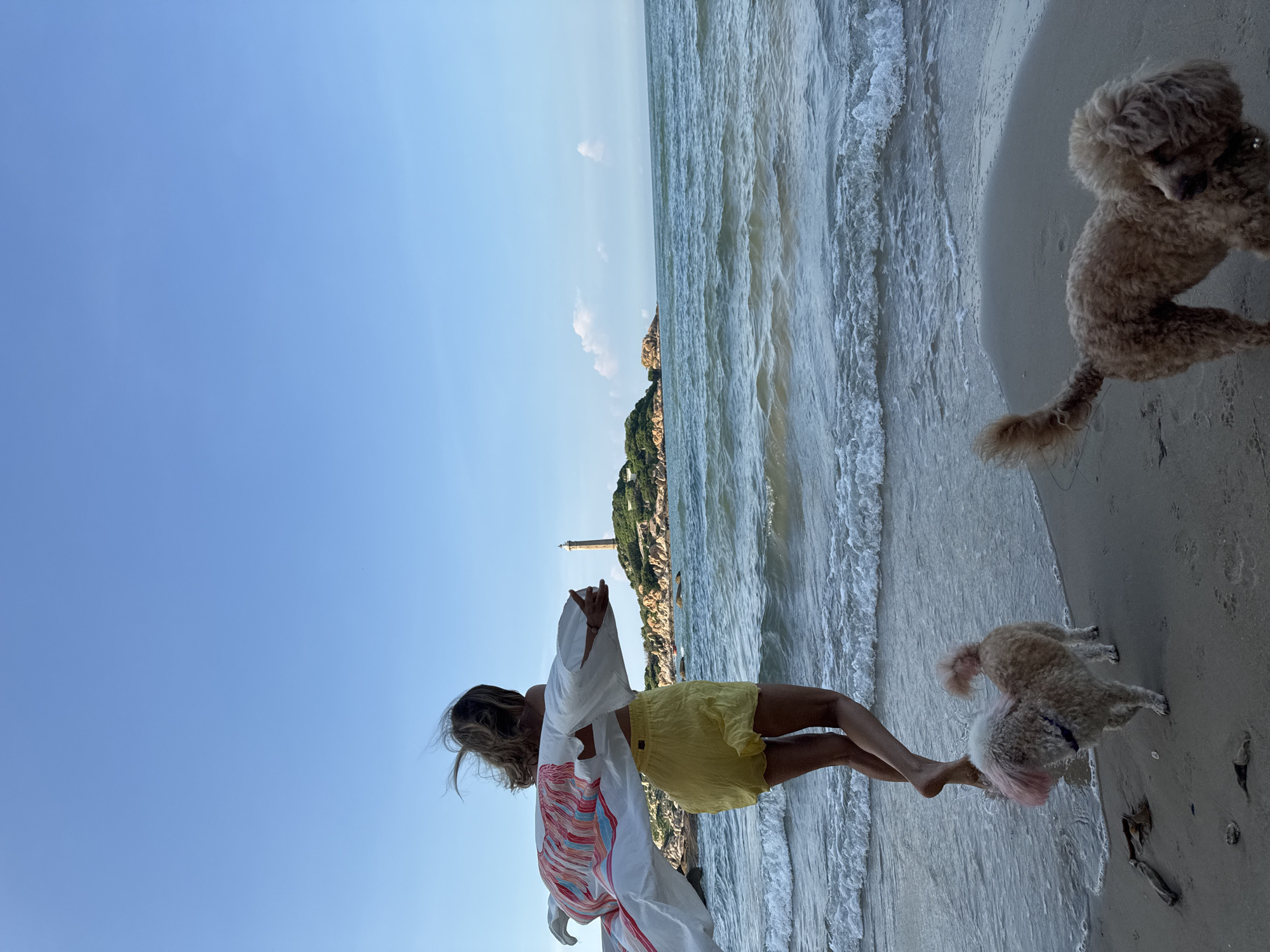 A woman with windblown hair, wearing a white patterned kimono and yellow shorts, stands on a sandy beach facing a lighthouse on a rocky island. Two dogs, one light brown with pink dyed fur and the other a tan poodle, are on the beach near her. Gentle waves lap the shore under a clear blue sky. The scene appears to be at Mũi Kê Gà, a peninsula in Thuận Thành, Vietnam.