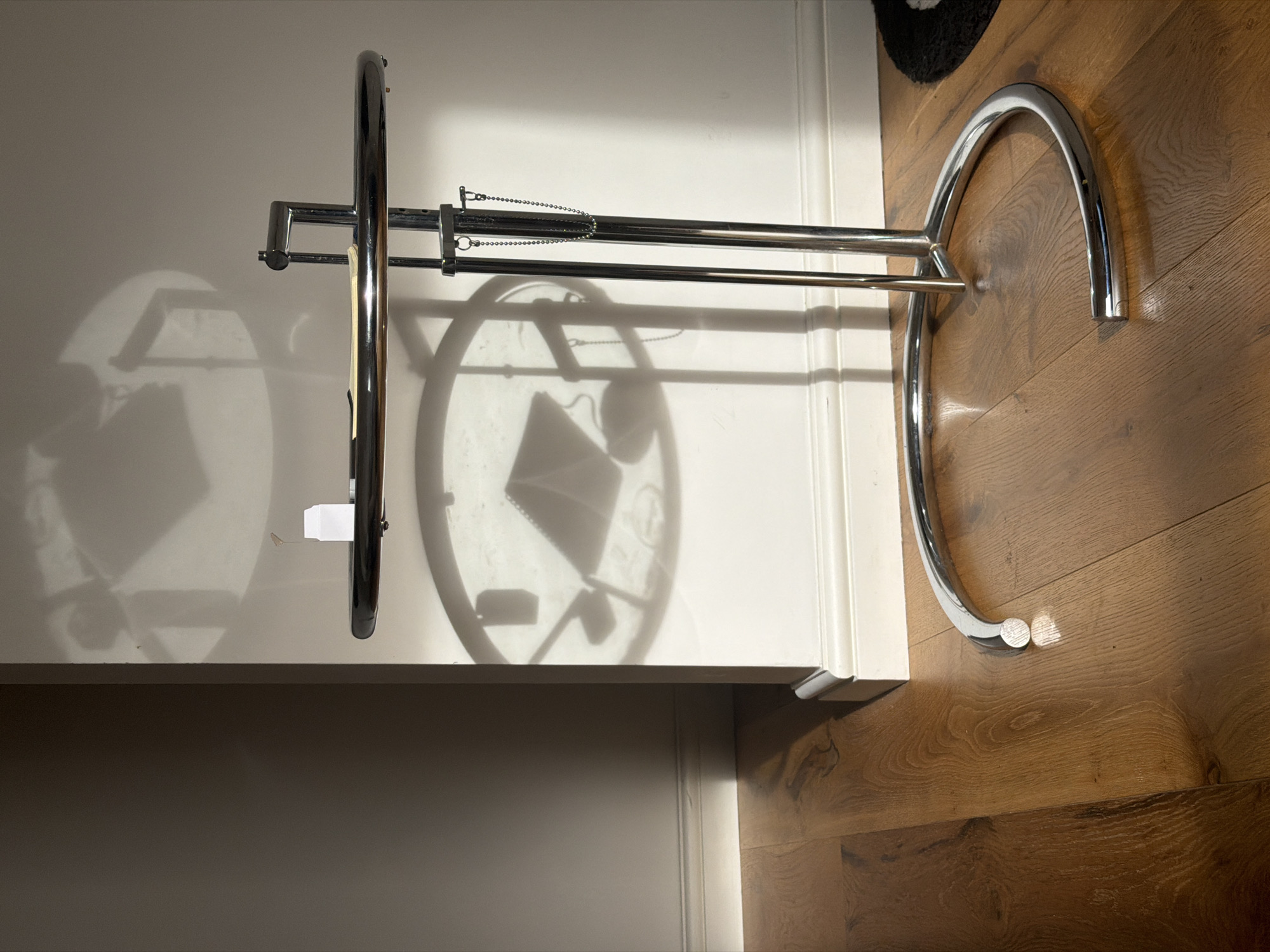 The image shows a chrome garment rack. It is positioned on a light-colored floor next to what appears to be a white cabinet. The lighting creates strong shadows on the cabinet's surface. The surrounding environment includes light-colored walls. The location is not discernible from the given image.