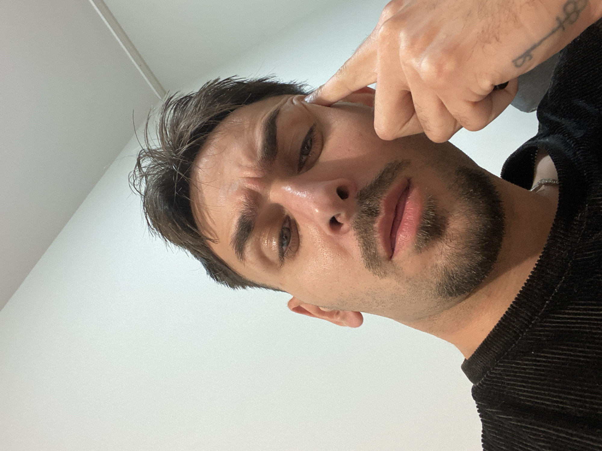 A close-up portrait shows a man looking directly at the viewer with an intense expression. His head is tilted, and his right index finger is pressed into or near his right ear, while his eyebrows are slightly furrowed. He has dark, somewhat messy hair, a short beard, and a mustache. His skin appears slightly shiny, possibly from oil or sweat.
He is wearing a black, ribbed or corduroy-like garment, and a silver chain necklace is visible around his neck. In the upper right background, his left hand is visible, adorned with a black ring on his ring finger. A small tattoo, appearing to be a stylized key or symbolic design, is visible on his left forearm or wrist. Due to the angle and clarity, any numbers or additional details of the tattoo are difficult to discern precisely (it resembles "4-8" or "18").
The background is a plain, light-colored (likely white or off-white) wall or ceiling, indicating an indoor setting. A thin, dark line, possibly a structural element or seam, is visible in the upper part of the background. The lighting is bright and even. No text is visible in the image. There are no specific visual cues to indicate the location as Sofia, Bulgaria.