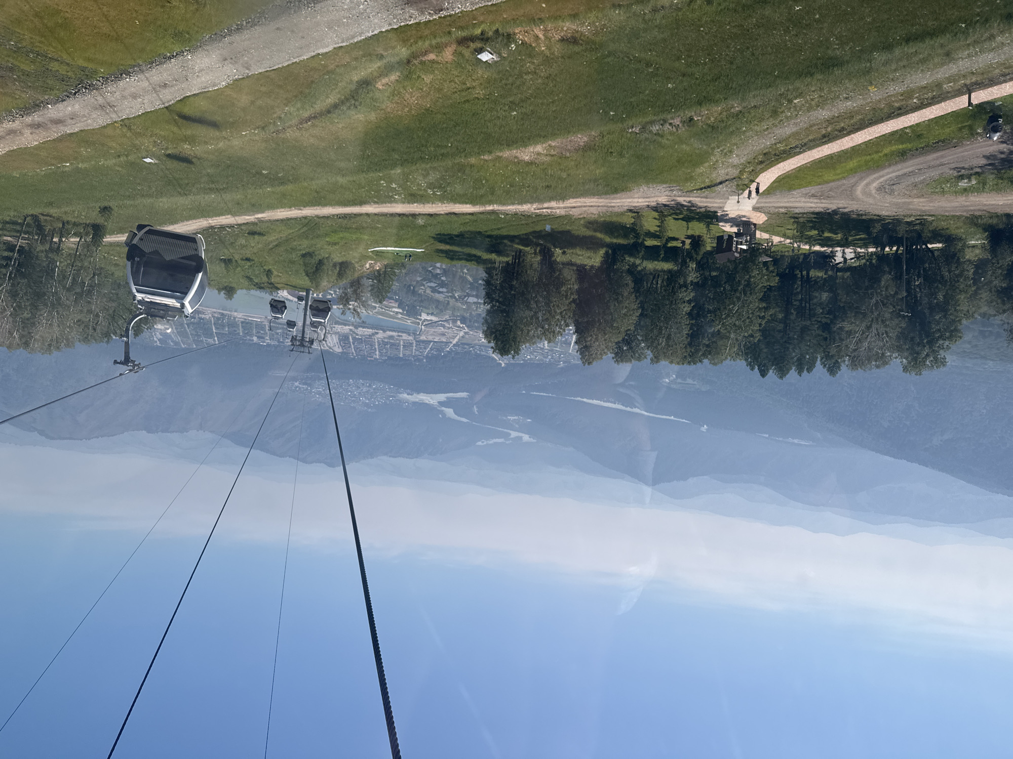 In Ozernoe, Russia, a cable car descends over a grassy mountainside. The cable car cabins are suspended from a thick wire cable, and several cabins are visible, moving towards the viewer. The landscape features rolling hills covered in green grass and scattered trees. In the distance, a hazy mountain range stretches across the horizon under a clear blue sky. A town is visible nestled in the valley below, with buildings and a river winding through it. The scene appears to be a popular tourist destination, offering panoramic views of the natural beauty and the settlement.