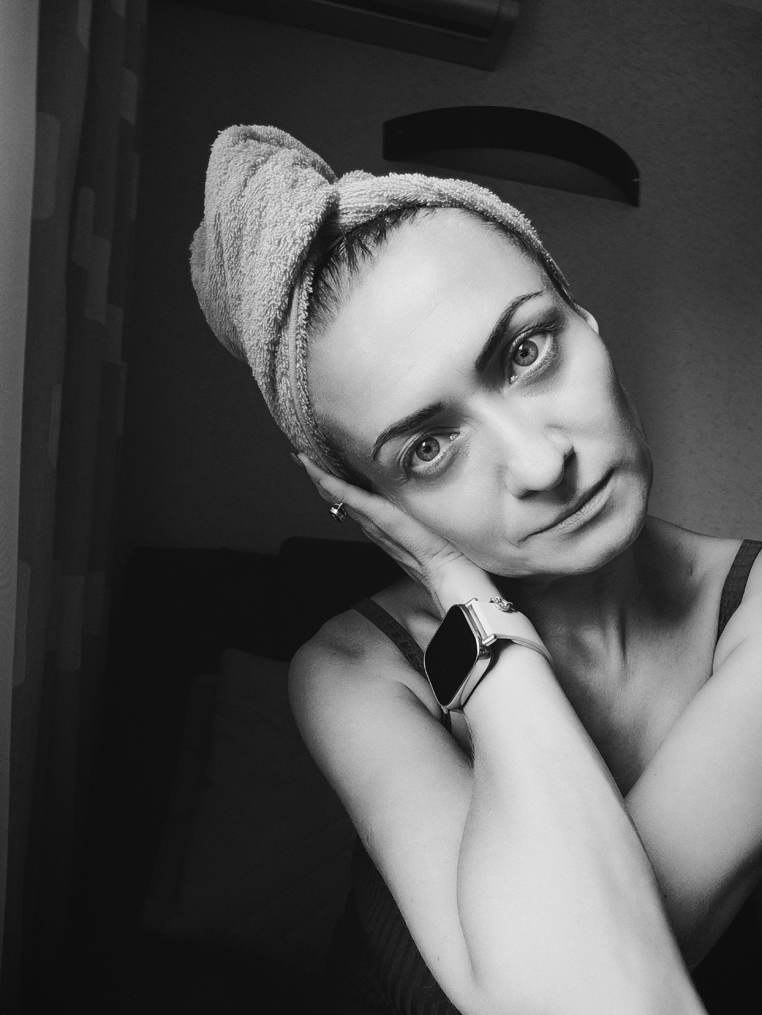 A black and white, medium shot portrait features a woman wearing a towel turban and a watch. She is leaning on her left hand, with her right arm crossed over her chest. Her head is tilted to the left, and she is looking directly at the camera. The background is dark and out of focus. The image appears to be taken indoors, possibly in a bathroom or bedroom. There is no indication of the location being Moscow, Russia.