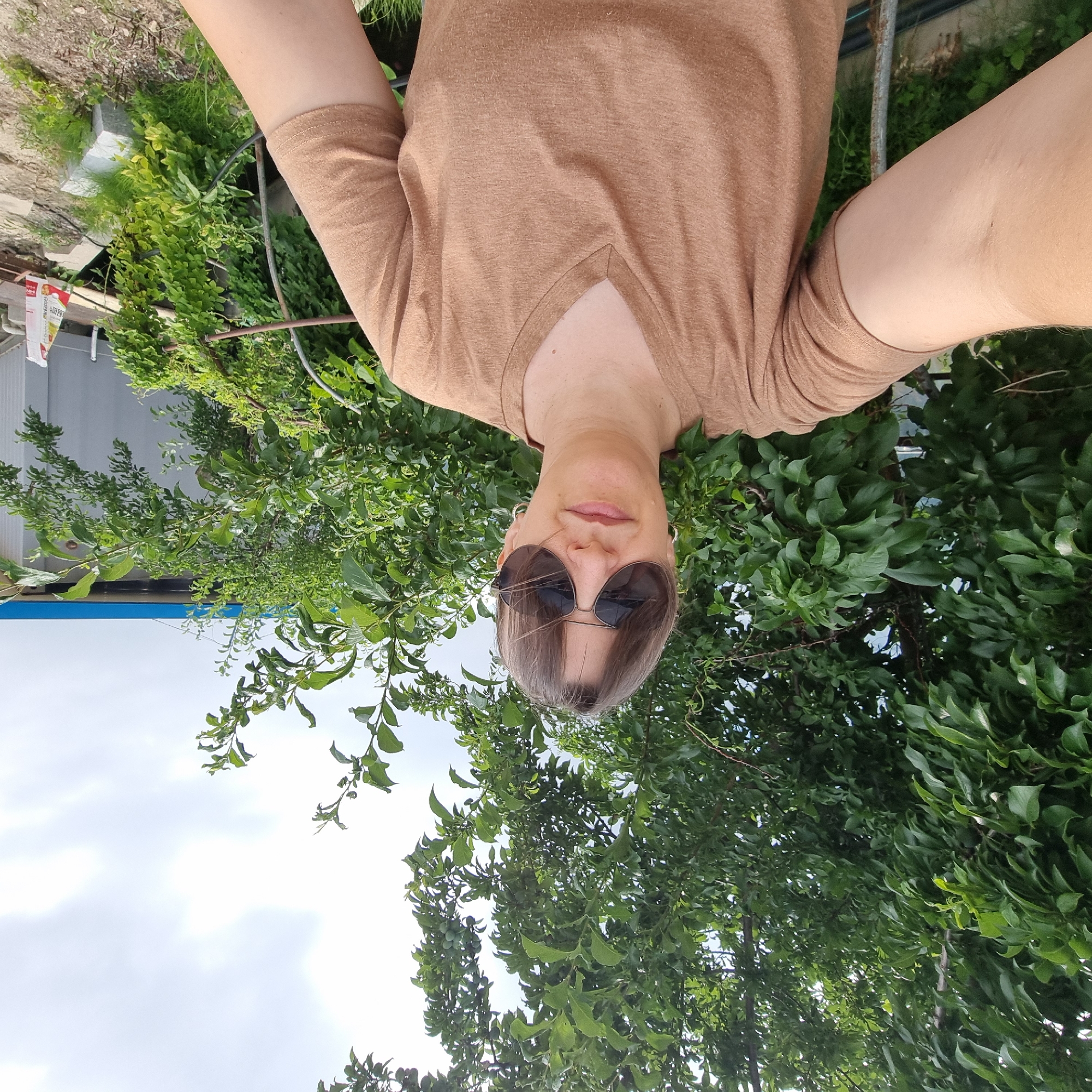 A woman with brown hair and sunglasses takes a selfie outdoors. She is wearing a brown v-neck t-shirt. She is standing in front of green trees, with a light blue building and some construction materials visible in the background. The location is Dunnae-myeon, South Korea.