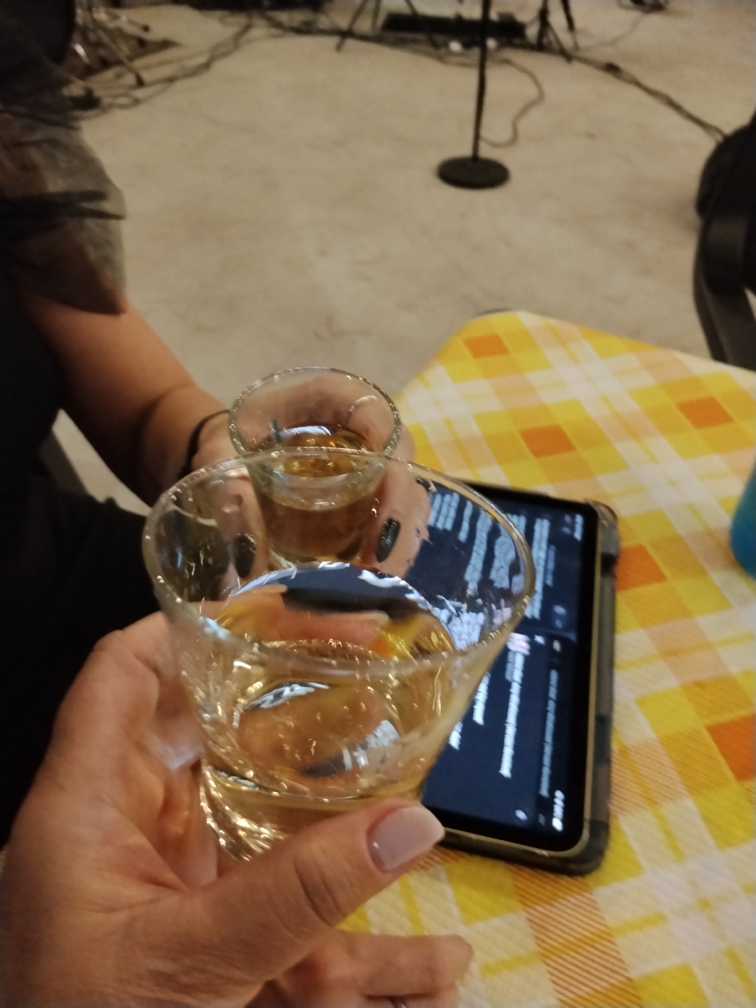Two hands are holding shot glasses filled with a clear amber liquid, clinking them together in a toast. The glasses are made of clear glass, and the liquid inside is translucent. One hand, on the left, is holding a larger, wider shot glass, while the other hand, further back and slightly to the right, is holding a smaller, taller shot glass. Both hands have well-maintained fingernails, with the visible fingernails on the left hand painted a light, neutral color. The background shows a beige, slightly textured floor, a black microphone stand with a round base, and some electrical cords. In the foreground, a portion of a tablet computer displaying text is visible, resting on a yellow and orange checkered tablecloth. The scene appears to be indoors, possibly in a bar or a casual gathering space. There is no visual information available to determine the specific location within the city of Toritto, Italy.