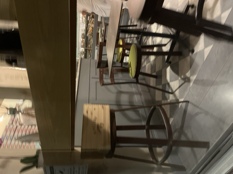 This image captures the interior of what appears to be a cafe or small eatery in Fiesole, Italy. The scene is static, showing an arrangement of furniture and a display area, possibly during quiet hours.
In the foreground and midground, several tables and chairs are visible. Some chairs feature dark frames with distinctive circular cushions in a bright chartreuse or lime green color, while others are simpler dark wooden chairs. A tall, dark-framed bar stool with a circular base is prominent on the right side. On the floor nearby, a light-colored wooden crate is present, displaying faint, unreadable text. The floor features a pattern of light and dark grey checkered tiles, which transitions into a plain light grey section. Shadows cast by the furniture are visible on the tiled floor.
In the background, a long counter or display case extends across the width of the room. Behind its glass, various items, likely food products such as pastries or packaged goods, are arranged. A person wearing a white top, possibly a staff uniform, is partially visible behind the counter. The overall lighting is artificial and somewhat subdued, contributing to a calm atmosphere. A large, light-colored vertical beam or wall element occupies the left side of the frame, with reflections of stacked items visible beyond it. A green plant in a pot is barely visible in the bottom left corner. No discernible activity is taking place. Apart from the faint text on the crate, no other text is clearly legible, including on a partially visible sign at the very bottom left.