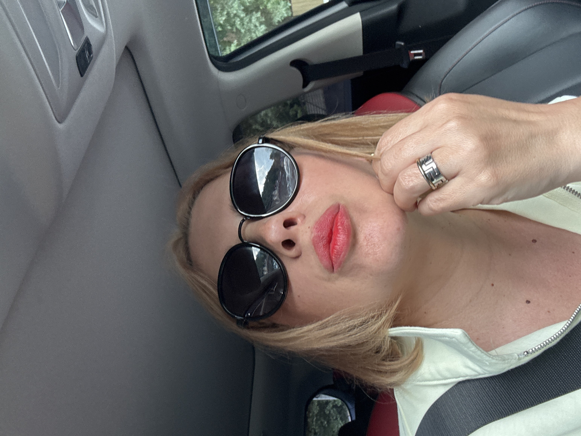 A woman with blonde hair and red lipstick is taking a selfie inside a car. She is wearing large, round, black sunglasses and a light-colored jacket. She is wearing a ring on her right hand, which is resting on her chin. The interior of the car, including a black leather seat and a seatbelt, is visible. The scene appears to be set in a city environment with trees and buildings visible through the car window.