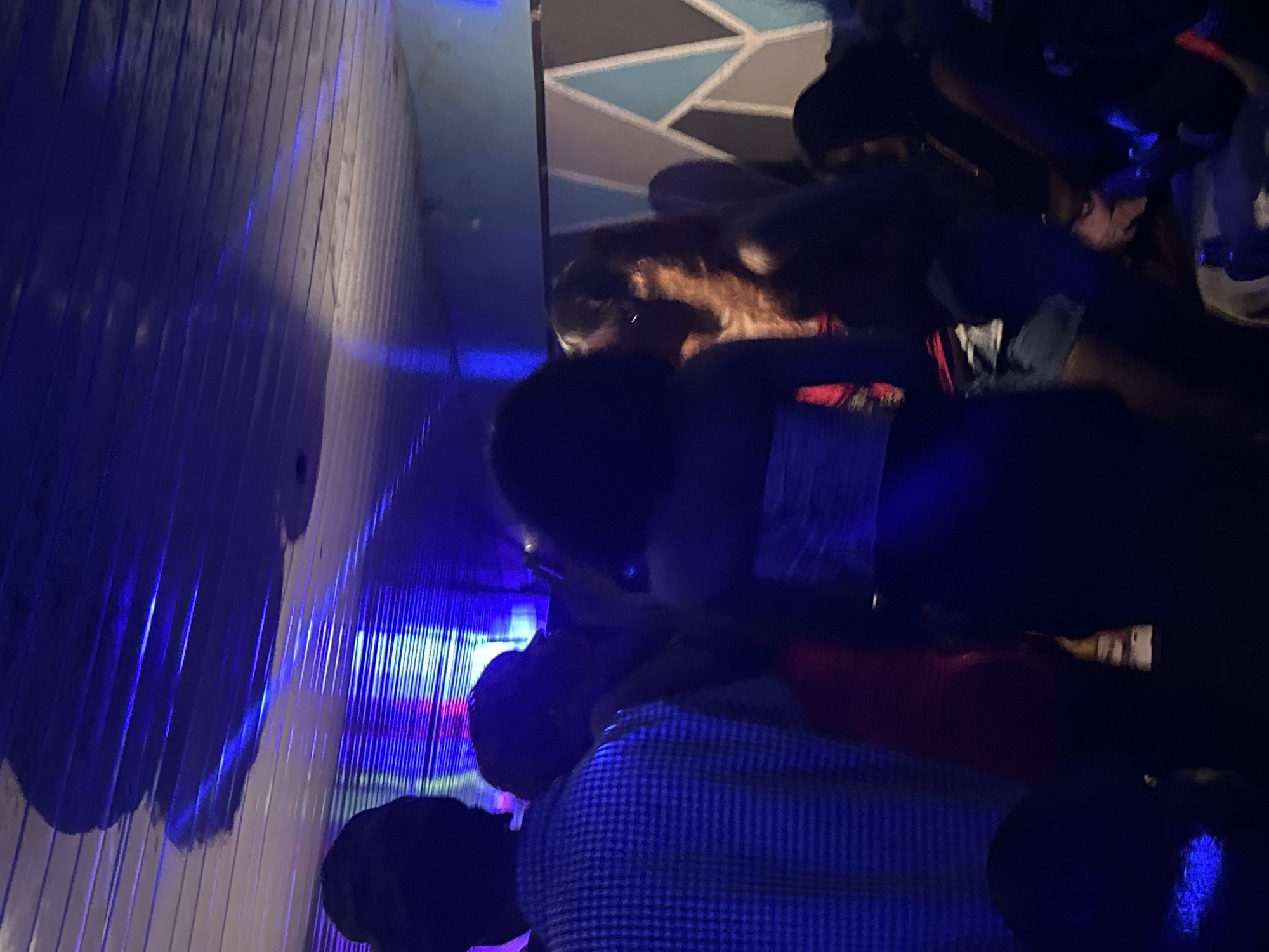 The image depicts an indoor scene, likely a nightclub or event space, illuminated by blue and purple lights. In the foreground, a person from the shoulders up is visible on the left, wearing a blue and white checkered shirt. To their right, a woman with dark, curly hair is seen from the back, wearing a white strapless top. Further to the right, another woman with long, light brown hair is partially visible, facing towards the left. The background features a wall with a geometric pattern of light blue, grey, and white shapes. The ceiling appears to be ribbed and reflects the ambient lighting. The overall atmosphere suggests a social gathering with people interacting. The location is identified as Wumat, Nigeria.