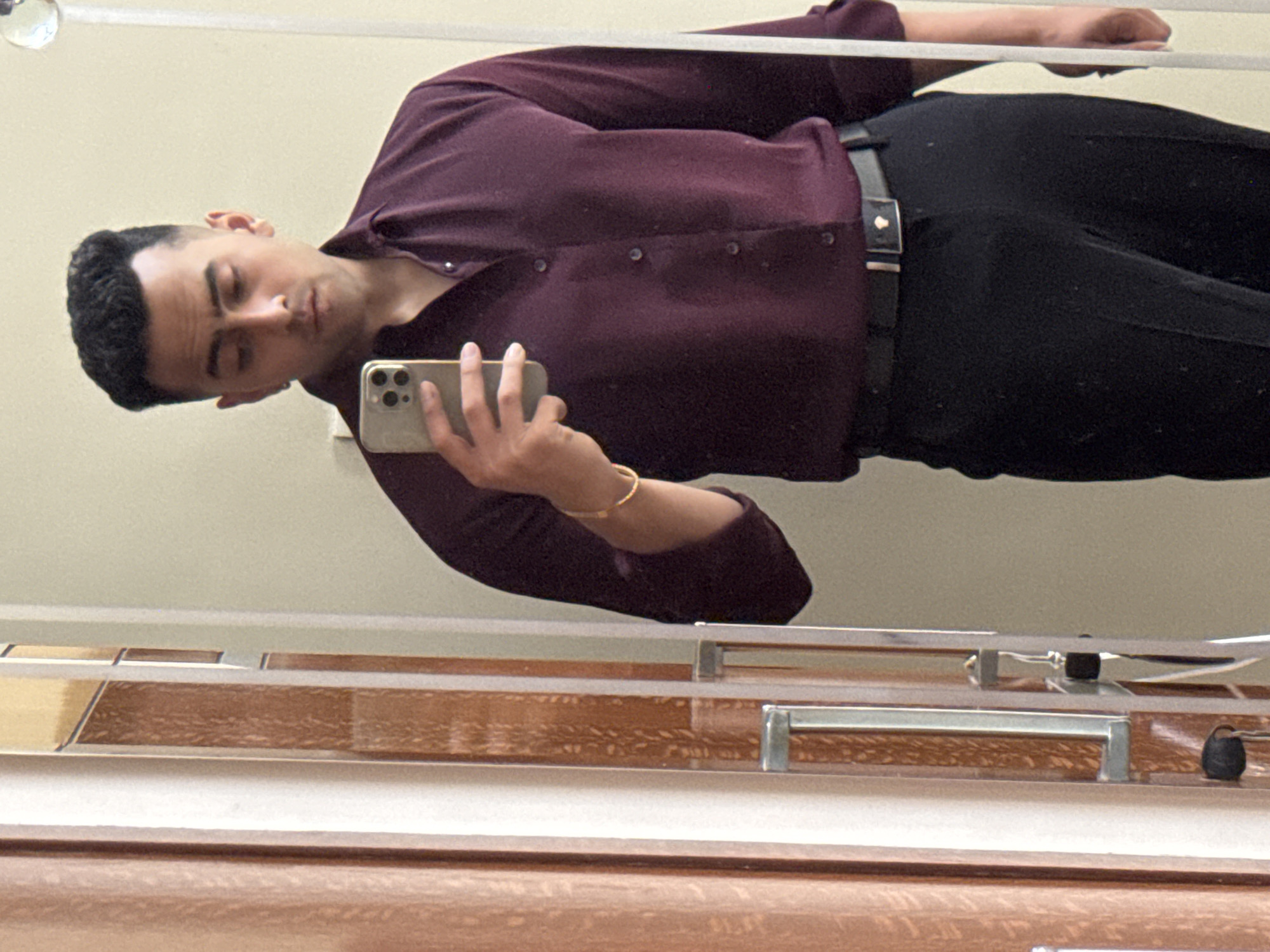 A man is taking a selfie in a mirror. He has short dark hair and is wearing a maroon button-down shirt and black pants with a black belt. He is holding a gold-colored smartphone in his right hand. The background is a cream-colored wall above a mirror frame with brown and silver accents. The location and city cannot be ascertained from the image alone.