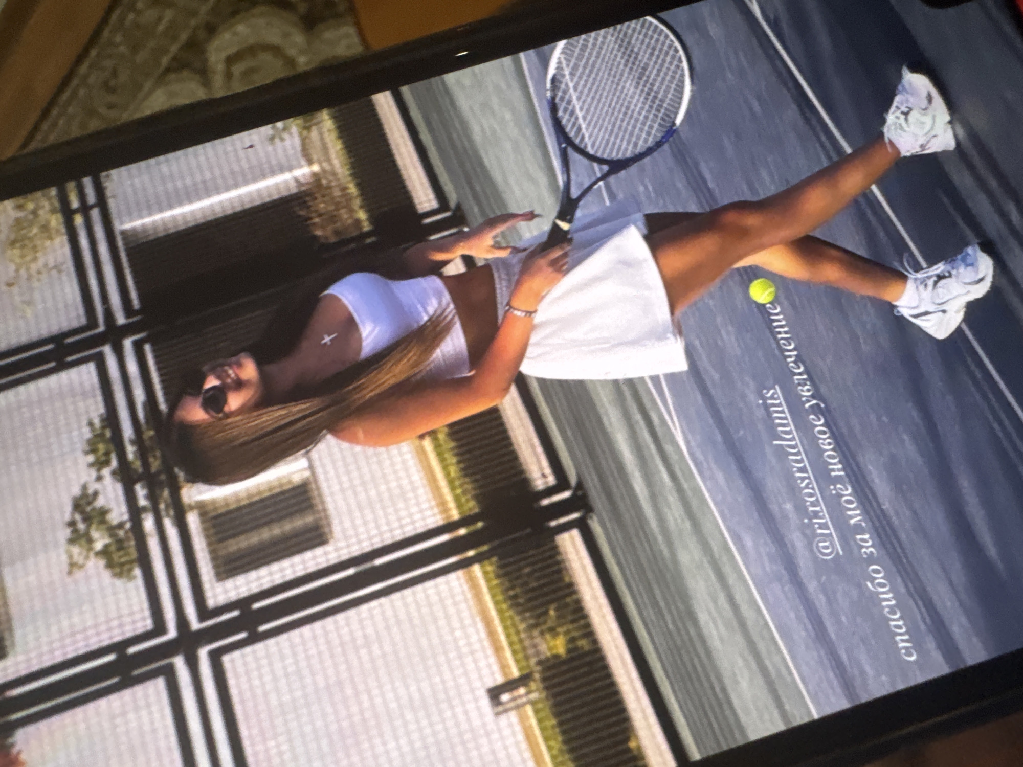 The image is a photograph of a screen displaying a vertically oriented picture of a woman on a tennis court. The screen itself is tilted, showing parts of the surrounding room with a patterned surface.
The primary subject is a woman with long, straight, dark hair, wearing sunglasses, a white halter-neck crop top, a white skirt or shorts, and white sneakers. She is standing with one leg bent and the other extended, posing on a dark-colored tennis court with white lines. She holds a tennis racket in her right hand, resting its head on the court. A yellow tennis ball is visible near her left leg. A small white cross pendant hangs around her neck, and she wears a bracelet on her left wrist. Her facial expression shows a slight smile.
The setting is an outdoor tennis court, indicated by the dark playing surface and white boundary lines. The background shows fencing and some greenery, suggesting an outdoor facility. The lighting appears to be bright and natural, consistent with daytime.
Text is visible on the tennis court surface within the displayed image. It is written in Russian and reads:
- "@ri.rosradamis" (likely an Instagram handle or tag)
- "спасибо за моё новое увлечение" (which translates to "thank you for my new hobby" or "thank you for my new passion").
The activity depicted is posing on a tennis court, suggesting an association with the sport, perhaps celebrating a new interest or a lifestyle image. The image quality from being a photo of a screen exhibits some grid patterns and slight blurriness.