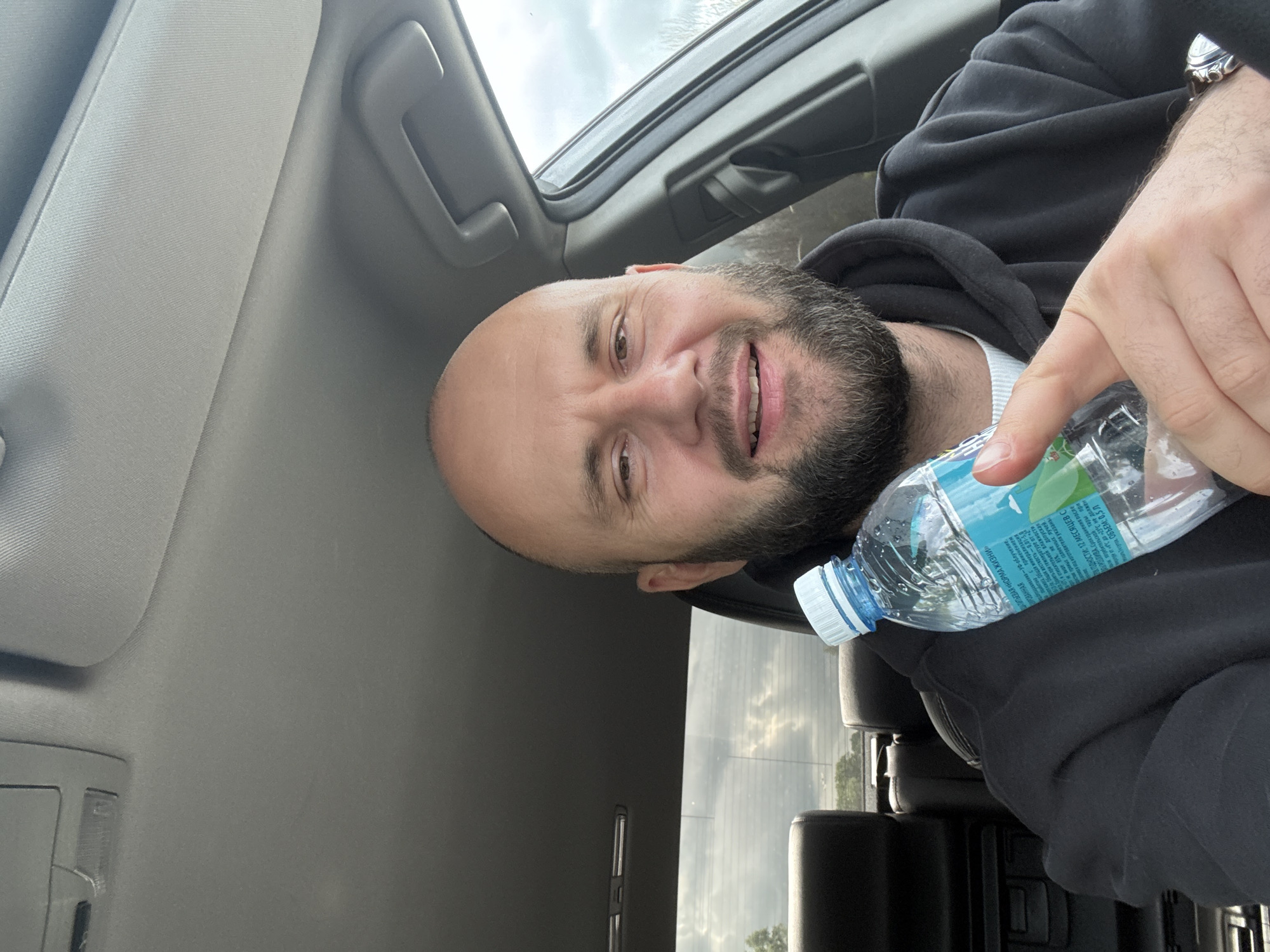 A man with a bald head and a beard is sitting in a car, holding a water bottle. The car's sunroof is open, and the sky is visible. He is wearing a black hooded sweatshirt. The interior of the car is grey. The label on the water bottle has text in Russian.