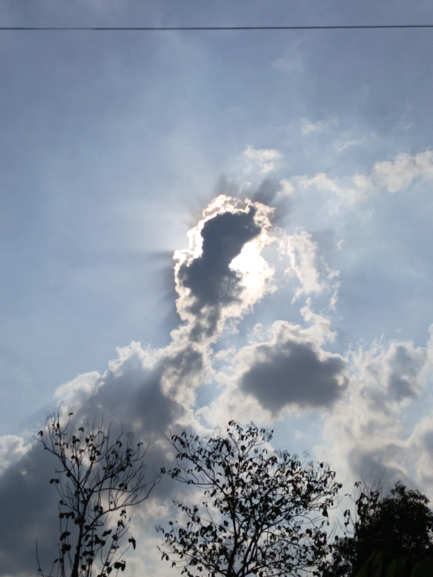 The image shows a cloudy sky with the sun partially obscured by a dark cloud, casting rays of light. The foreground features silhouetted trees. The scene is identified as being in Gedung Nyapah, Indonesia. There are no people or actions depicted.