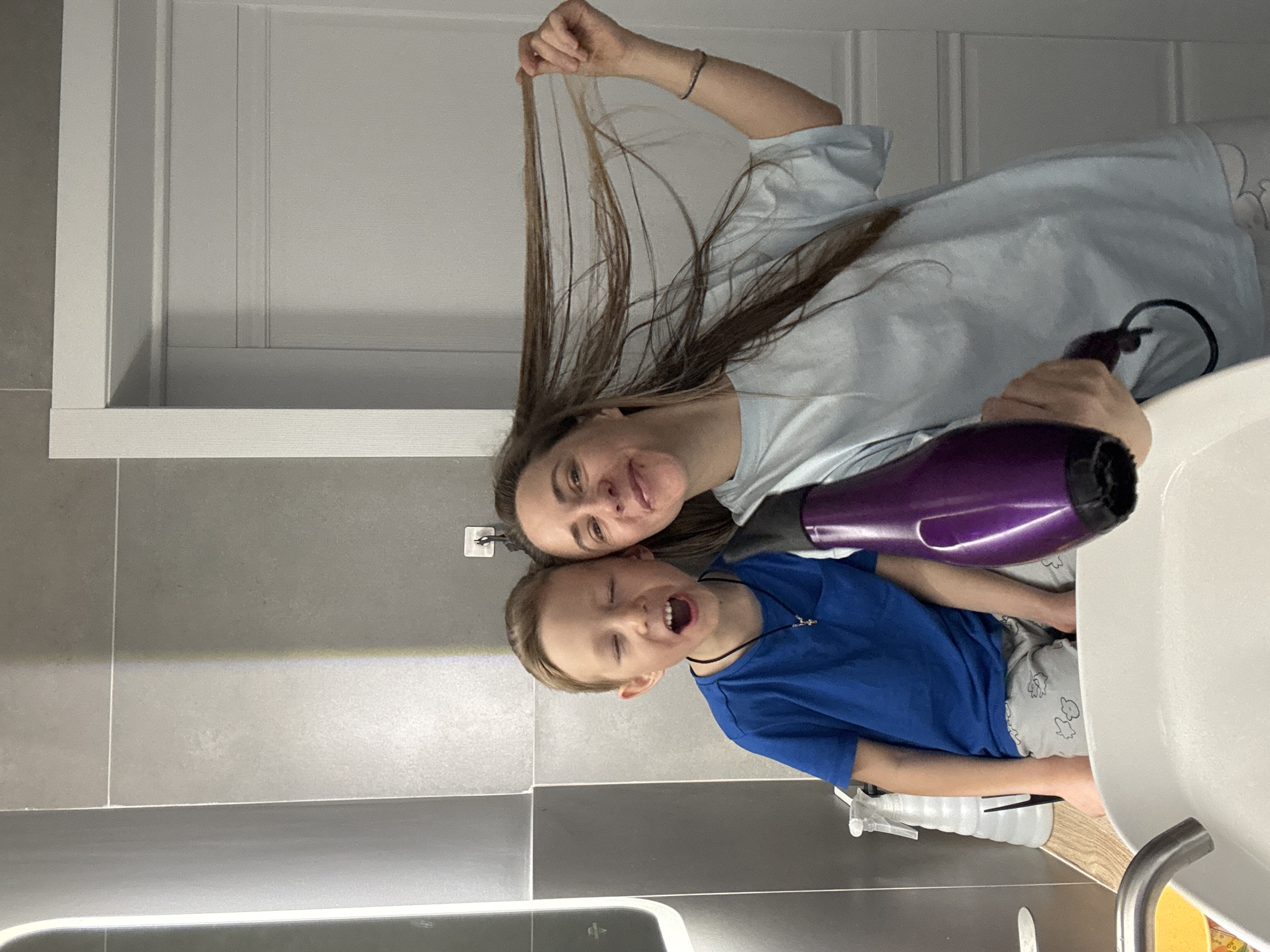 A woman and a young boy are in a bathroom. The woman is holding up a strand of her brown hair and using a purple hairdryer. The boy is next to her, smiling. The bathroom has gray tile walls and a white vanity with a sink. The location is likely Chaikovsky, Russia.