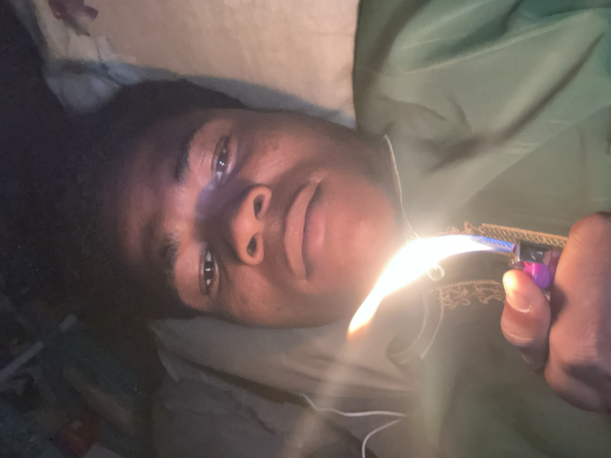 This media file captures a close-up of a person lying down, with their head and upper torso visible. The person has dark skin and dark hair. They are wearing a green garment that appears to be a shirt or robe, with some decorative trim visible. The person's right hand is holding a pink and purple lighter, which is ignited and emitting a bright flame. The flame is partially obscuring the person's face, casting a strong light on their features. The background is soft and out of focus, suggesting an indoor setting, possibly a bedroom. The lighting is dim, with the exception of the bright flame from the lighter. The overall mood of the image is intimate and perhaps a bit contemplative. No text is visible in the image. Given the location context of Ihiagwa, Nigeria, the scene likely takes place in a residential area. The time of day is not explicitly indicated, but the dim lighting could suggest evening or nighttime.