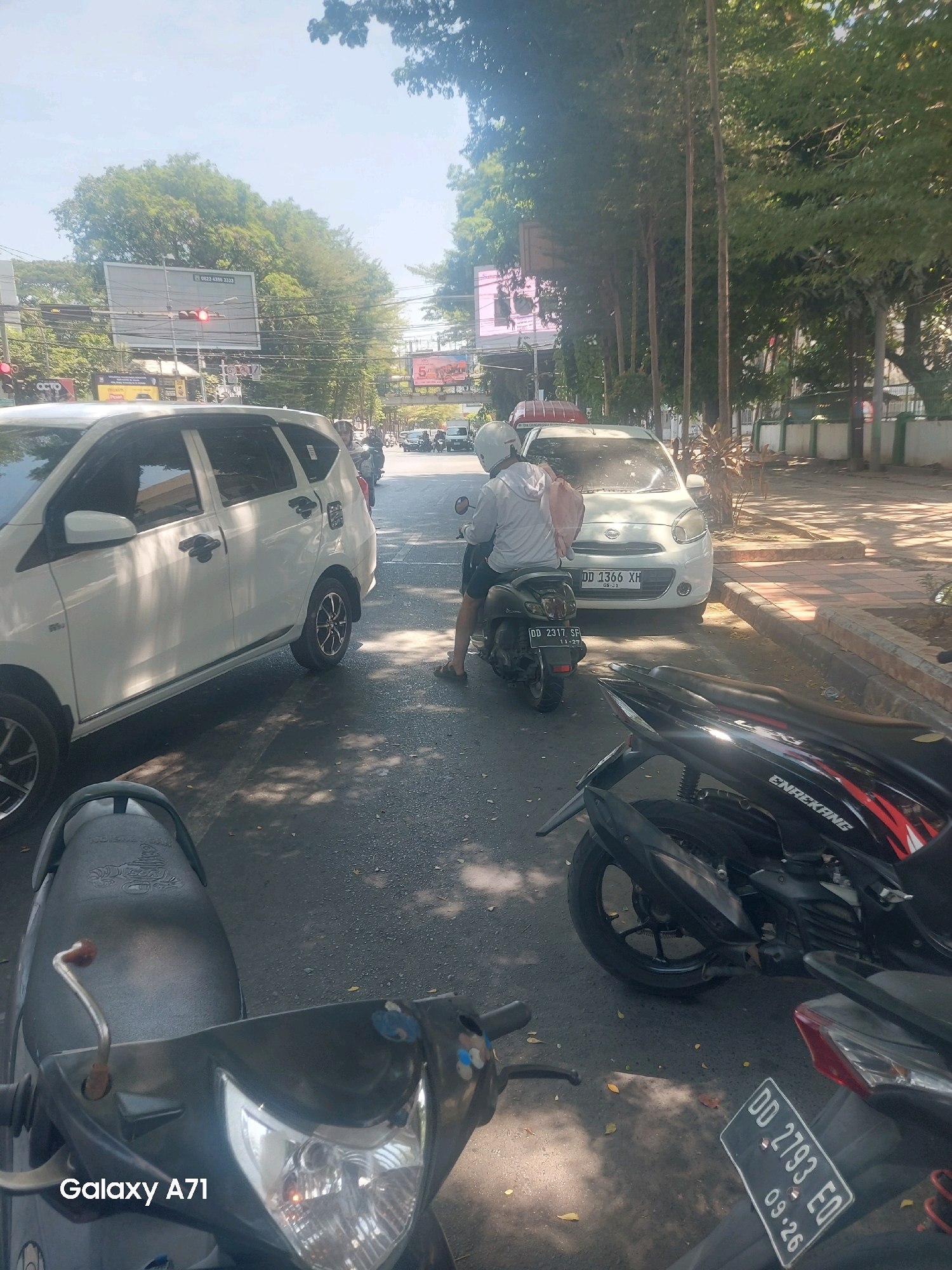 This is a street-level photograph taken in Makassar, Indonesia, on a sunny day. The scene depicts a traffic jam on a paved road lined with trees. In the foreground, two motorcycles are parked, one partially obscuring the other. The motorcycle on the left has a dark seat and a visible "Galaxy A71" watermark. The motorcycle on the right is black with red and white graphics and a license plate that reads "DD 2793 EQ" and "09.26".
Further down the road, a white sedan with license plate "DD 1366 XH" is visible. To the left of the sedan, another white vehicle, possibly an SUV or minivan, is also part of the traffic. A person wearing a white helmet and a light-colored hoodie is riding a dark-colored scooter in the middle of the traffic lane, carrying a pink backpack. Several other vehicles and pedestrians are present further in the background. A pedestrian bridge spans the road in the distance. Billboard advertisements are visible on the left side of the street, one of which displays a red traffic light. The overall atmosphere suggests a busy urban environment during daylight hours.