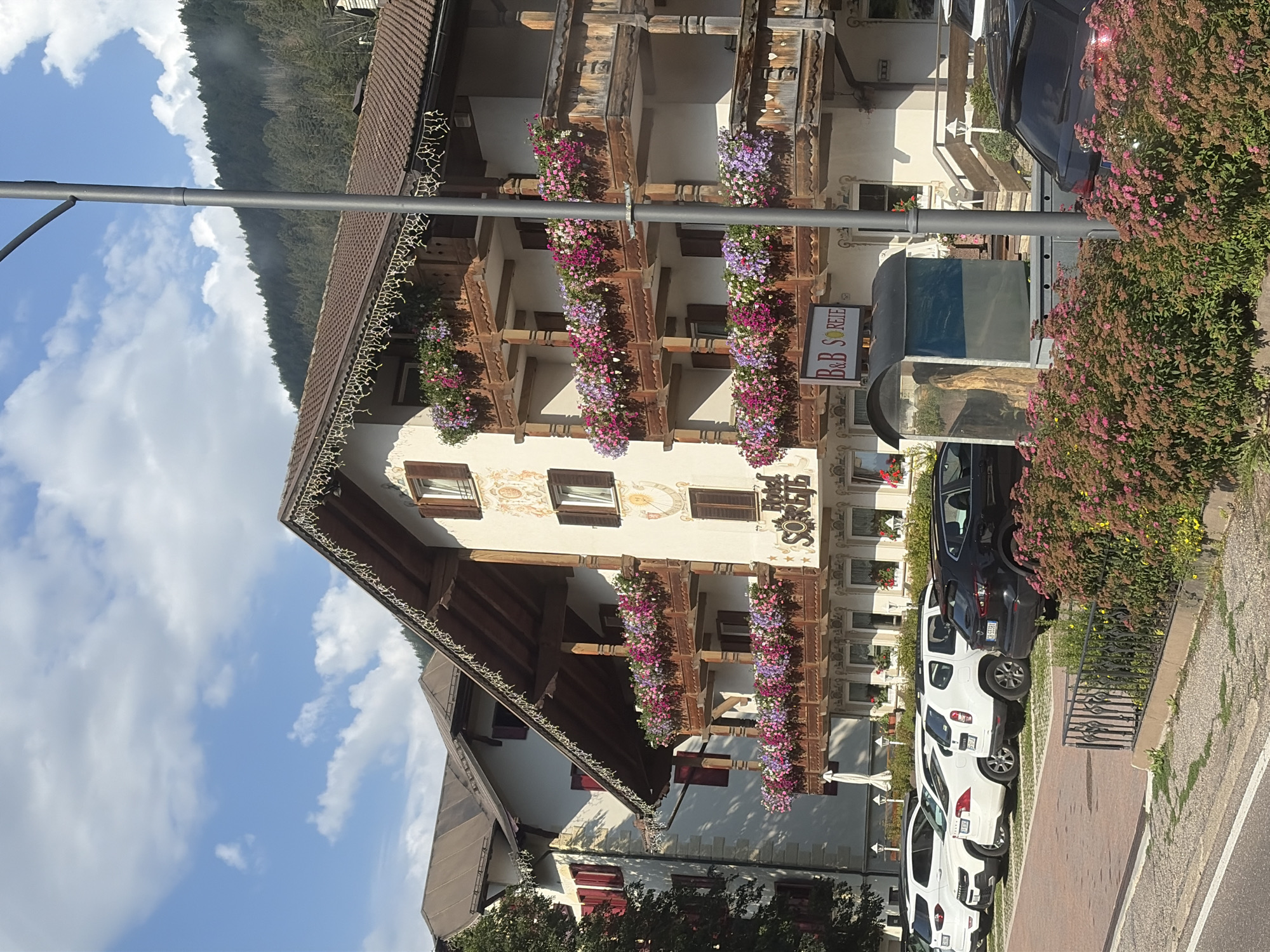 The image shows a hotel and B&B named "B&B SOREIE" in Pozza di Fassa, Italy. The hotel is a multi-story building with traditional alpine architecture, featuring wooden balconies adorned with numerous colorful flowers. The building has a brown tiled roof with decorative trim. A parking lot is visible in front of the hotel, with several cars parked, including white SUVs, a white sedan, and a black SUV. A lamppost stands in front of the hotel. The background shows a dense forest covering a mountain slope under a blue sky with white clouds. The scene appears to be captured during daytime.
