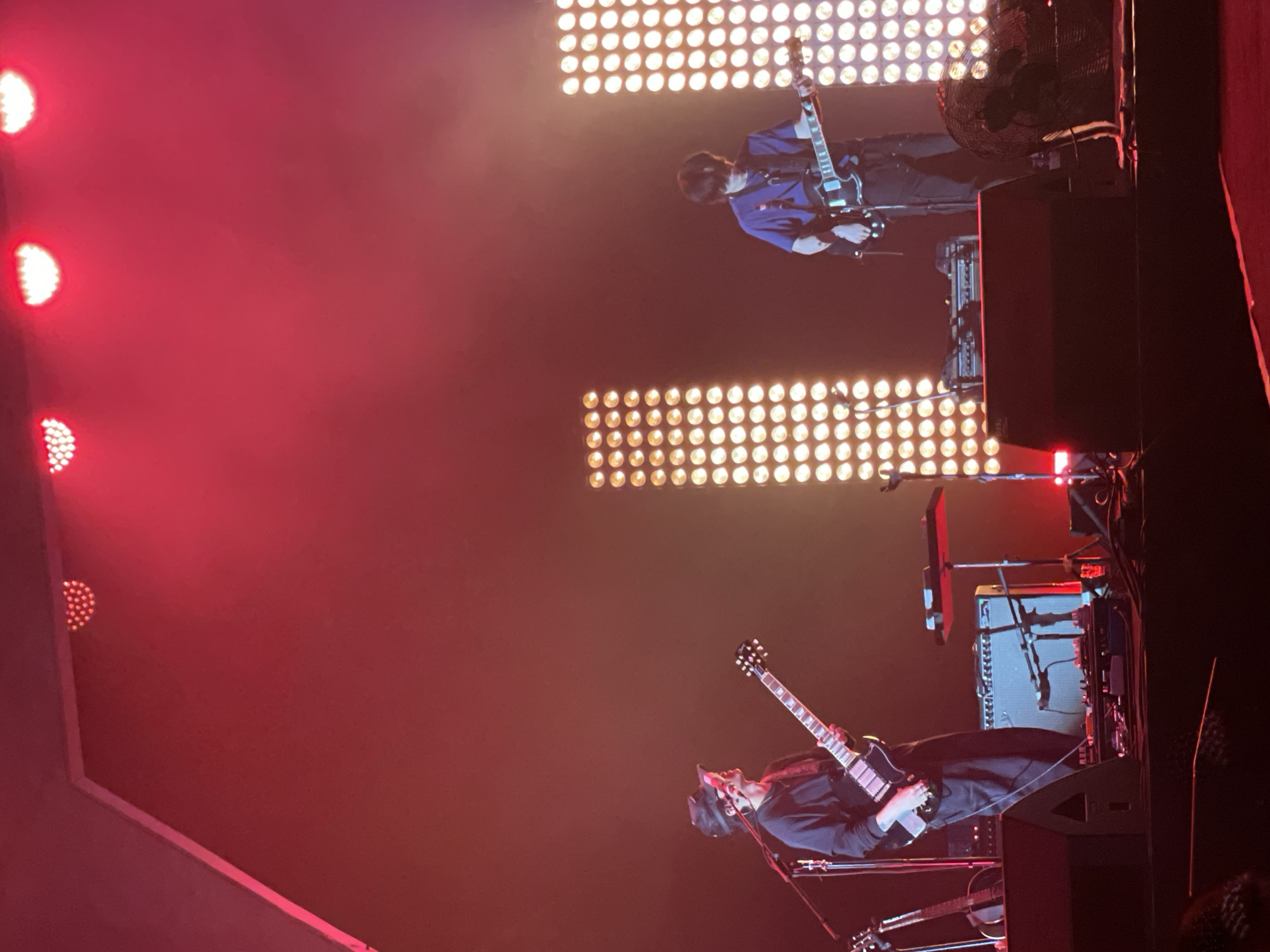 The image captures a music performance in Salembaran, Indonesia. Two guitarists are visible on stage, illuminated by a combination of red and white stage lights. One guitarist is wearing a blue jacket and playing a light-colored electric guitar, while the other is dressed in black and playing a dark electric guitar. Stage equipment, including amplifiers and a fan, is visible behind the musicians. The overall environment suggests a live concert setting.