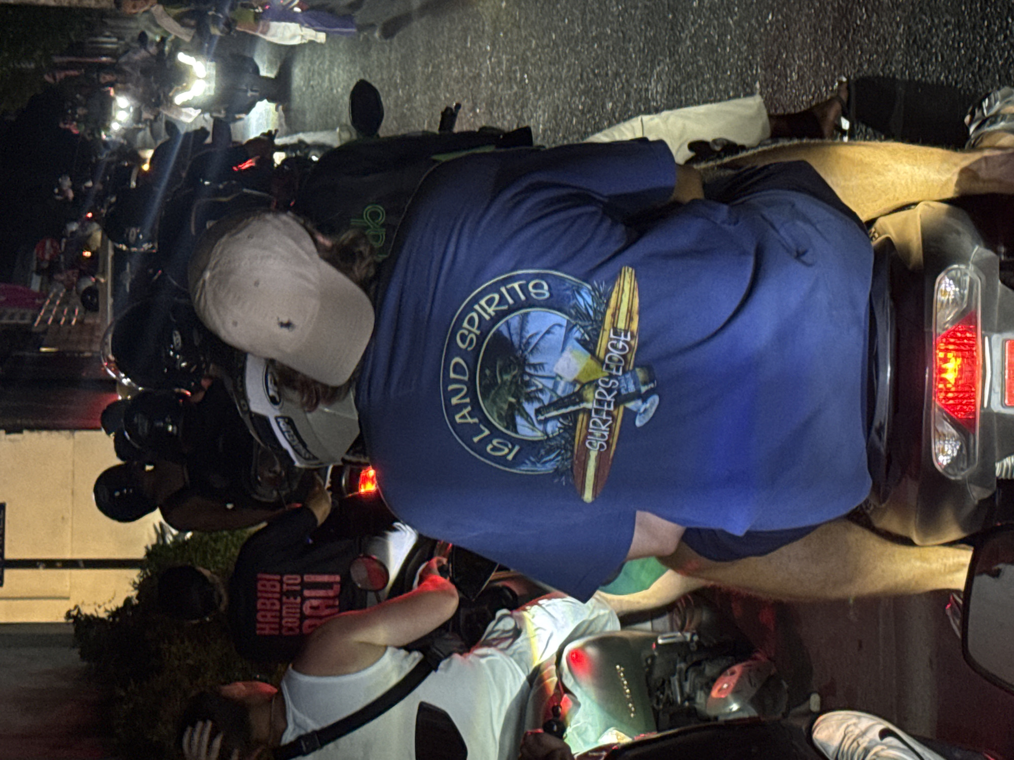 The image captures a crowded street scene at night, possibly in North Kuta, Indonesia. The scene is dominated by a gathering of motorcycles parked closely together. Several people are visible, some wearing helmets and riding motorcycles, others standing beside them. One person is wearing a blue t-shirt with a "Island Spirits Surfers Edge" logo, and another person is wearing a shirt that says "Habibi Come to Bali." The street appears to be illuminated by the headlights and taillights of the vehicles. The overall impression is one of a bustling urban gathering, possibly related to evening or nighttime activity.