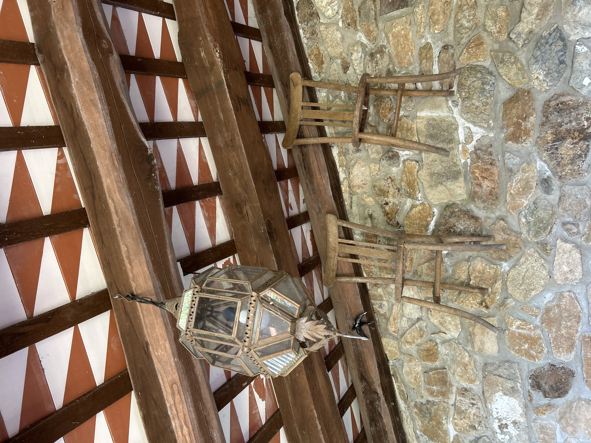 This image depicts an interior view looking upwards at a beamed ceiling and a stone wall. The ceiling is constructed of exposed wooden beams supporting a decorative pattern of alternating red and white triangular tiles. Hanging from one of the beams is an ornate, multi-faceted lantern made of glass and metal.
Attached to the stone wall are two simple wooden chairs, oriented sideways and appearing to be hung as decorative elements rather than for seating. The stone wall itself is composed of irregularly shaped stones of various earthy tones, cemented together. A small, dark metal decorative bird is attached to one of the wooden beams, near the lantern.
The scene suggests a rustic or traditional architectural style, possibly a patio, covered walkway, or interior space in a building with a stone facade. The lighting appears natural, indicating it's daytime. There are no people visible, and the overall mood is static and decorative. The provided location context places this scene in Tordera, Spain. No visible text is present in the image.
