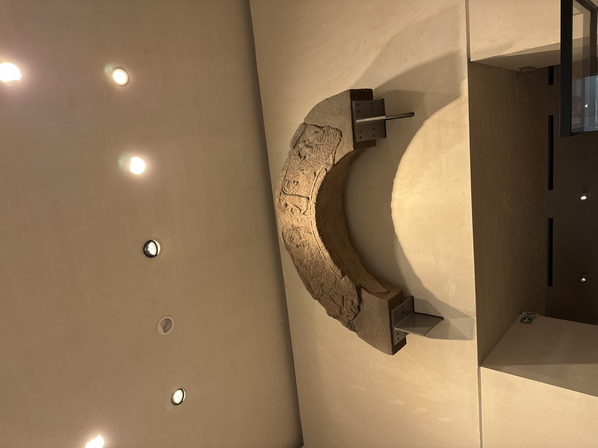 An ancient stone archway, carved with relief figures, is mounted on a cream-colored wall in a museum setting in the City of Edinburgh, United Kingdom. The arch is supported by two dark grey metal brackets attached to the wall. The ceiling above the arch features several recessed spotlights and one surface-mounted light fixture. The arch itself has a rough, weathered texture and displays faces with distinct features, as well as other carved designs that are partially obscured. Below the arch, a doorway or entrance is visible, with a cream-colored wall leading into a darker interior space. There are two green and white directional exit signs on the wall to the left of the opening. The overall lighting is dim, with spotlights creating distinct pools of light and shadow.