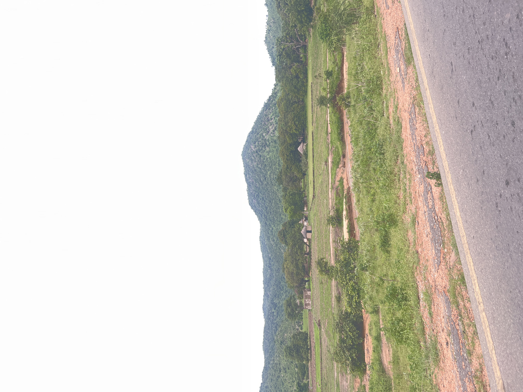 The image depicts a rural landscape in Minda, Nigeria. A paved road with a yellow line runs along the right side of the frame, bordered by a strip of dry, reddish-brown earth and sparse vegetation. To the left of the road is a large, grassy field that stretches towards a densely forested hill. The hill is covered in a variety of trees and appears to be the dominant geographical feature of the scene. In the mid-ground of the field, several small, thatched-roof structures, possibly huts, are visible, suggesting a small settlement. The overall impression is of a bright, clear day, given the abundant natural light and the lack of any visible clouds. The scene conveys a sense of peaceful, everyday rural life. There is no visible text in the image.