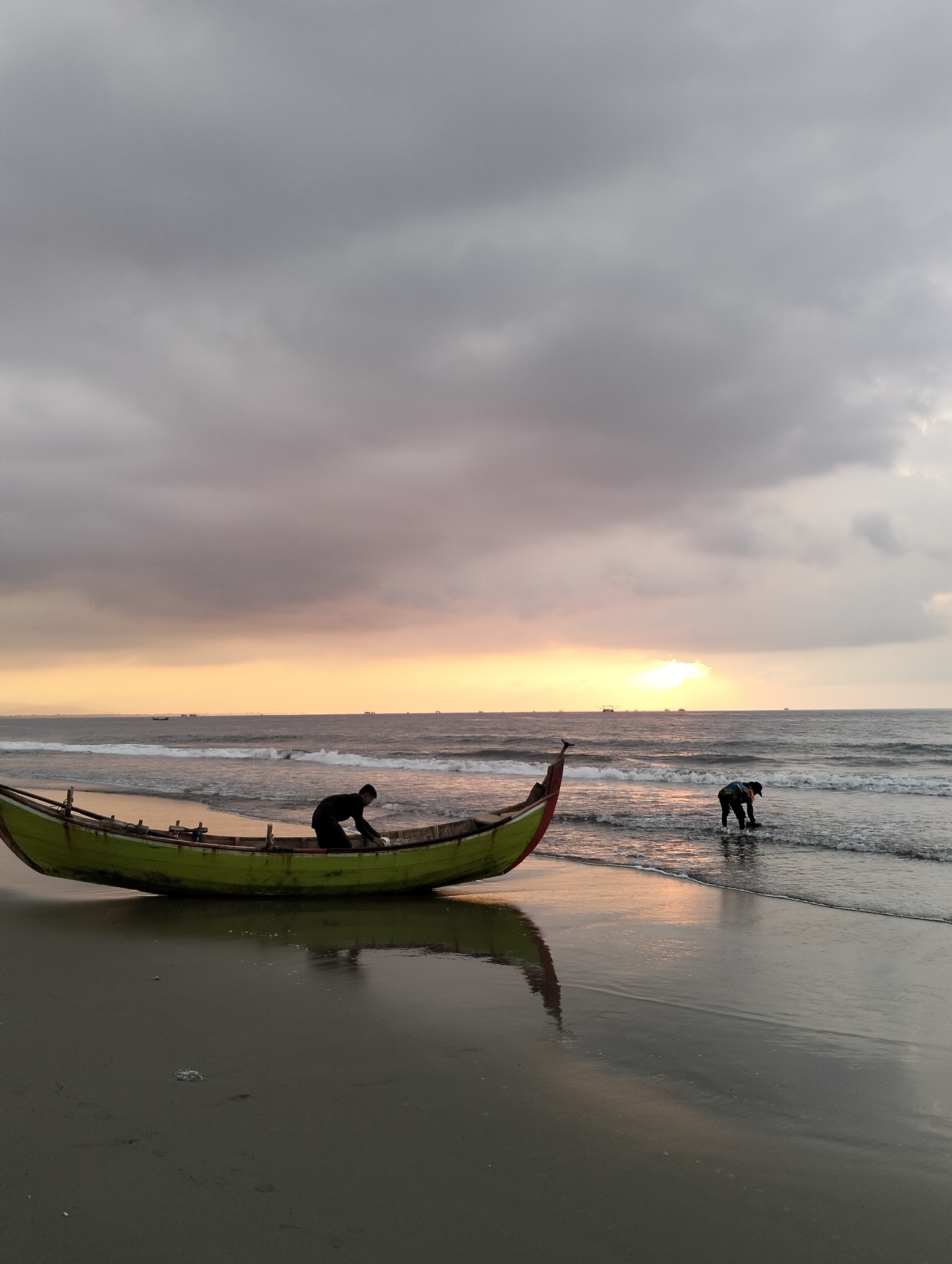 A wide-angle, eye-level shot captures a tranquil scene on the beach of Peuneulet Baroh, Indonesia, at dusk. A traditional, long, narrow fishing boat, painted green with a red accent on its stern, rests on the wet sand. The boat is positioned diagonally, with its bow facing left and its stern towards the ocean. Inside the boat, a person dressed in dark clothing is bent over, appearing to be cleaning or organizing something within the vessel.
To the right of the boat, the ocean stretches out, with moderate waves breaking gently on the shore, creating a foamy white line where the water meets the sand. In the shallow water, another person, also in dark clothing, is visible, facing away from the camera and appearing to be engaged in an activity near the water's edge. The sky above is dominated by a dramatic display of clouds, a mix of dark grays and lighter tones, with a warm, diffused orange glow emanating from the horizon, indicative of the setting sun. The light reflects on the wet sand and the surface of the ocean, creating a muted, atmospheric quality. In the distance, on the horizon, several small boats or ships are silhouetted against the sky. The overall impression is one of peaceful labor and the natural beauty of a coastal sunset.