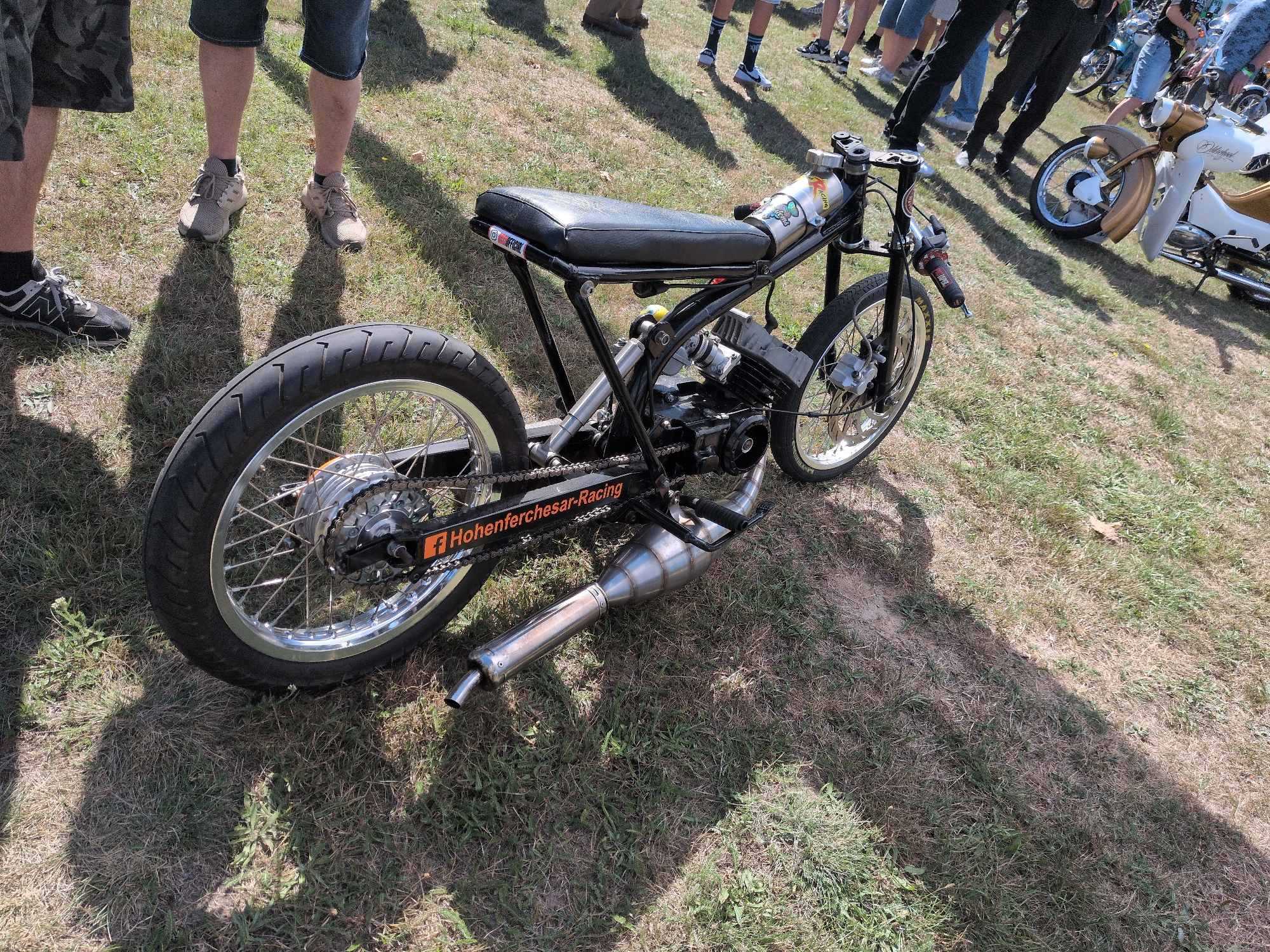 The image captures a custom-built motorcycle, possibly a moped, parked on grassy terrain. The motorcycle features a black seat, a prominent exhaust pipe, and a spoked rear wheel. A sticker on the chain guard reads "Hohenferchesar-Racing," indicating a potential connection to the town of Hohenferchesar, Germany. In the background, several individuals are visible, some standing and others appearing to be walking, all within an outdoor setting that suggests a gathering or event. The ground is covered with a mix of dry and green grass, with shadows cast by the sun, indicating it is daytime. The overall scene is consistent with an outdoor event or exhibition, possibly a motorcycle show or rally, taking place in or near Hohenferchesar.
