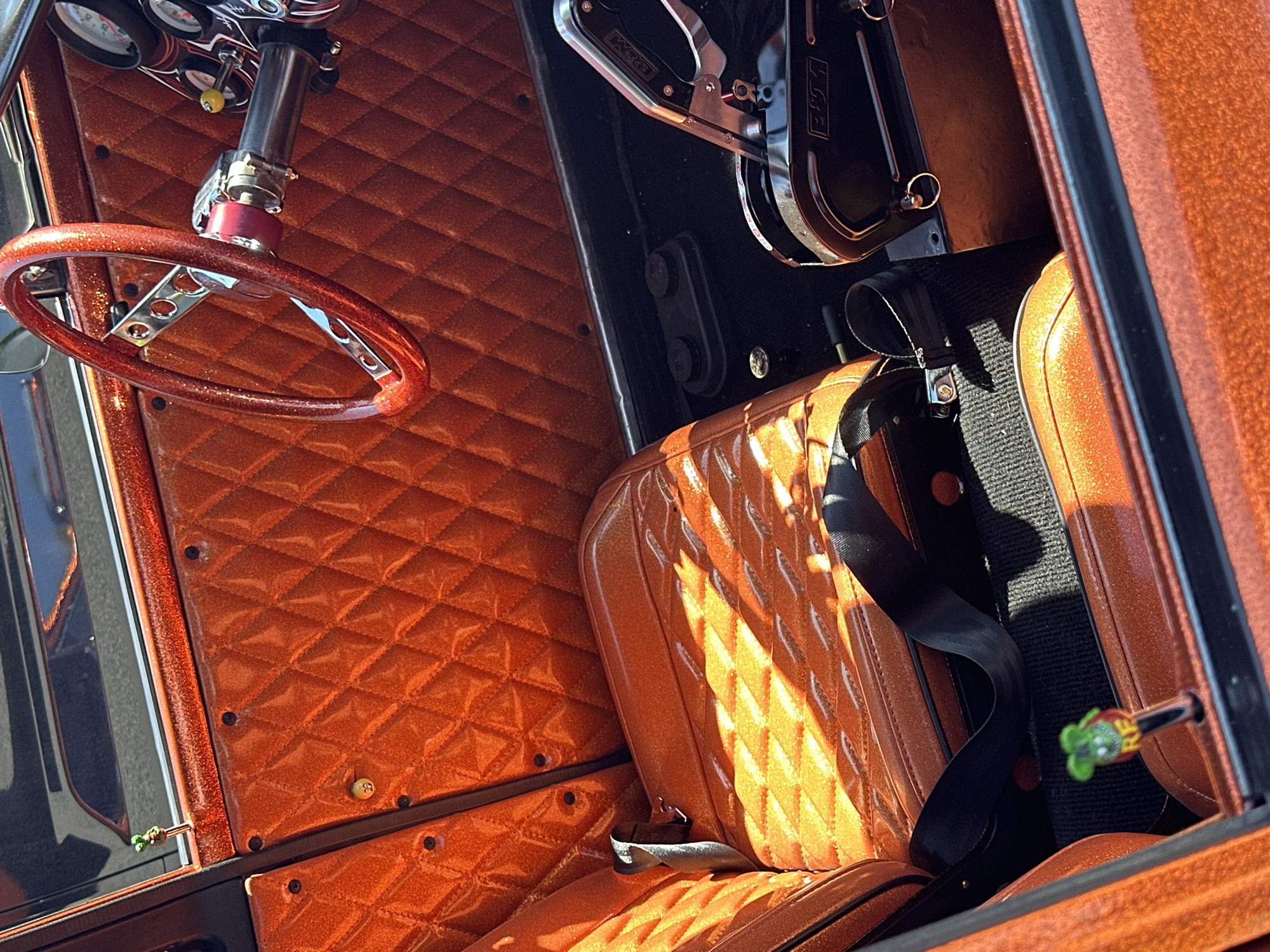 The image shows the interior of a vehicle, likely a custom car. The interior is predominantly orange with a diamond-stitched pattern on the seats and door panels. A red steering wheel is visible on the left side. The seats have black seatbelts. The vehicle's gear shift is located on the right. A small green-and-red object is protruding from the right door panel. The location, Port Moody, Canada, is not evident in the image.