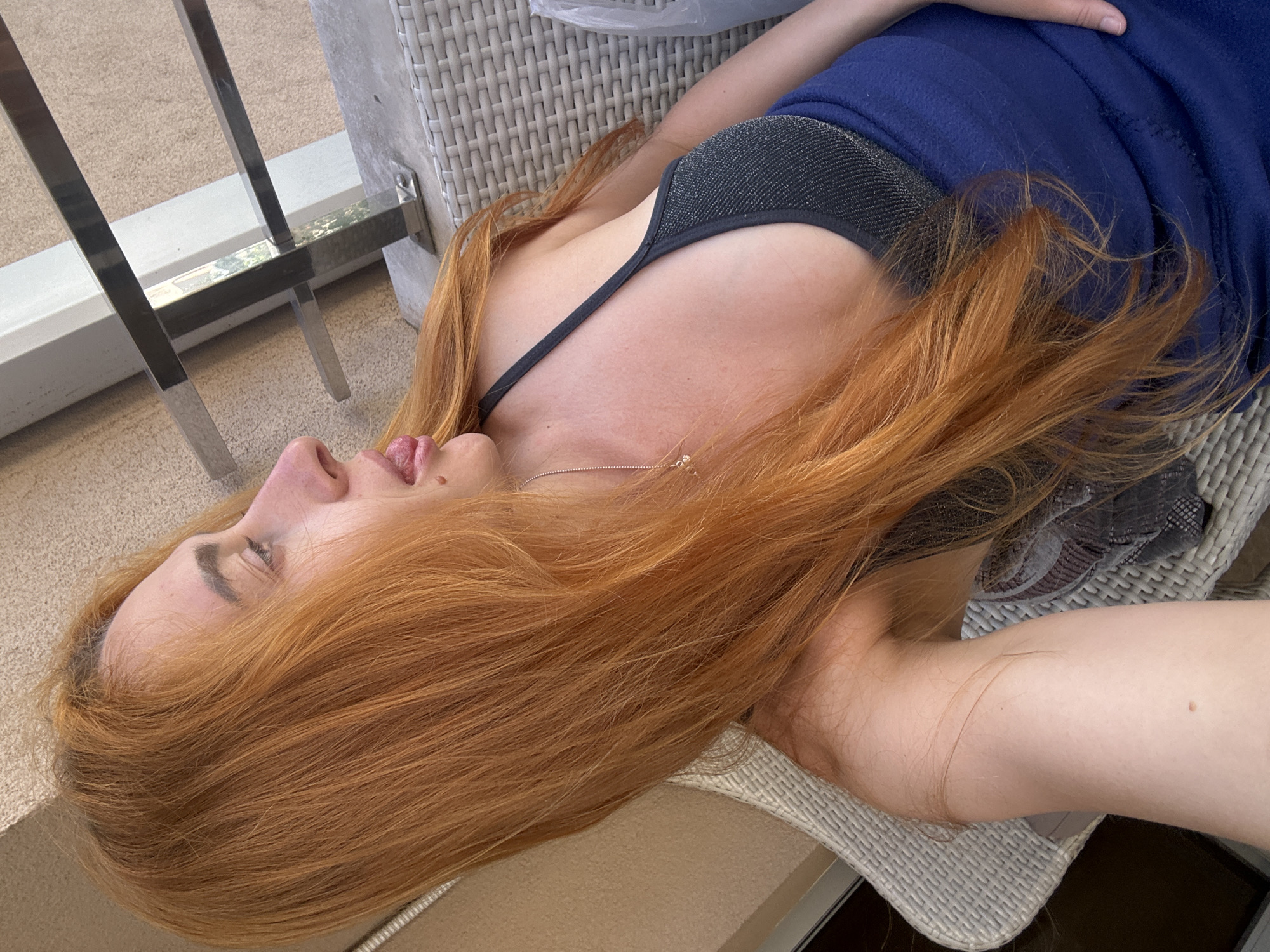 A woman with long, red hair is sitting on a wicker chair on a balcony, possibly in the city of Budva, Montenegro. She is wearing a dark top with a glittery texture and a blue garment. Her tongue is sticking out slightly. A metal railing is visible in the background, along with a beige wall. The lighting suggests it is daytime.