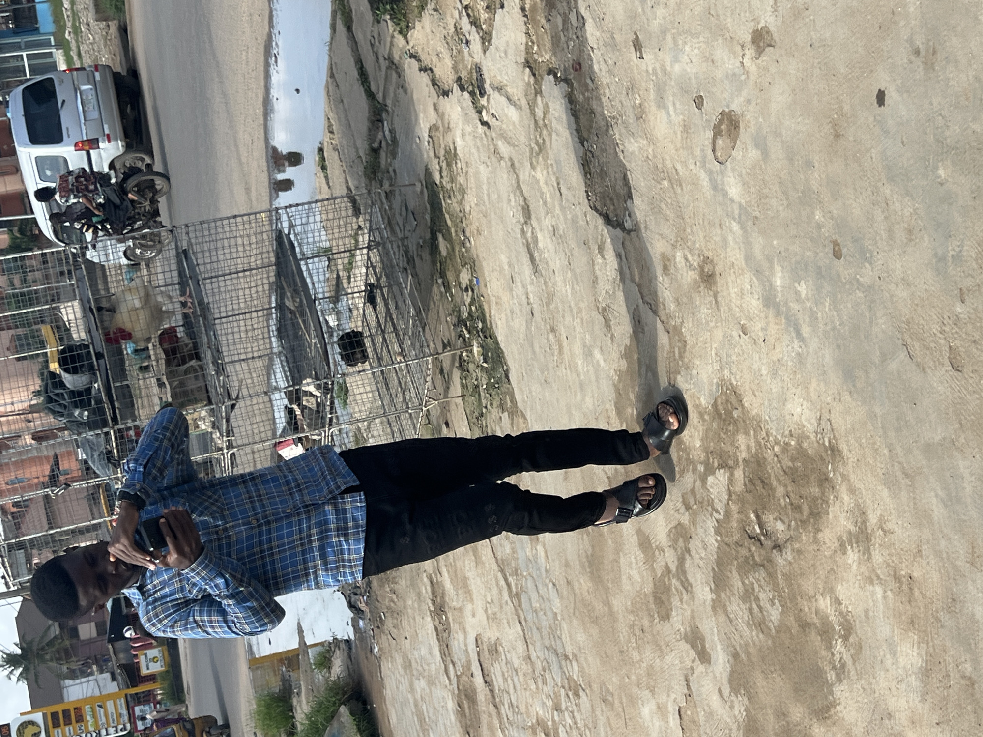 A man stands on a concrete surface in what appears to be an outdoor setting in Ijegun, Nigeria. He is wearing a blue plaid shirt, black pants, and black sandals. He is holding a mobile phone up to his face.
Behind him is a row of wire cages. Beyond the cages, there is a street with parked vehicles, including a white van and motorcycles. There are also buildings and trees visible in the background. The ground is cracked and discolored. A puddle of water is visible on the street.