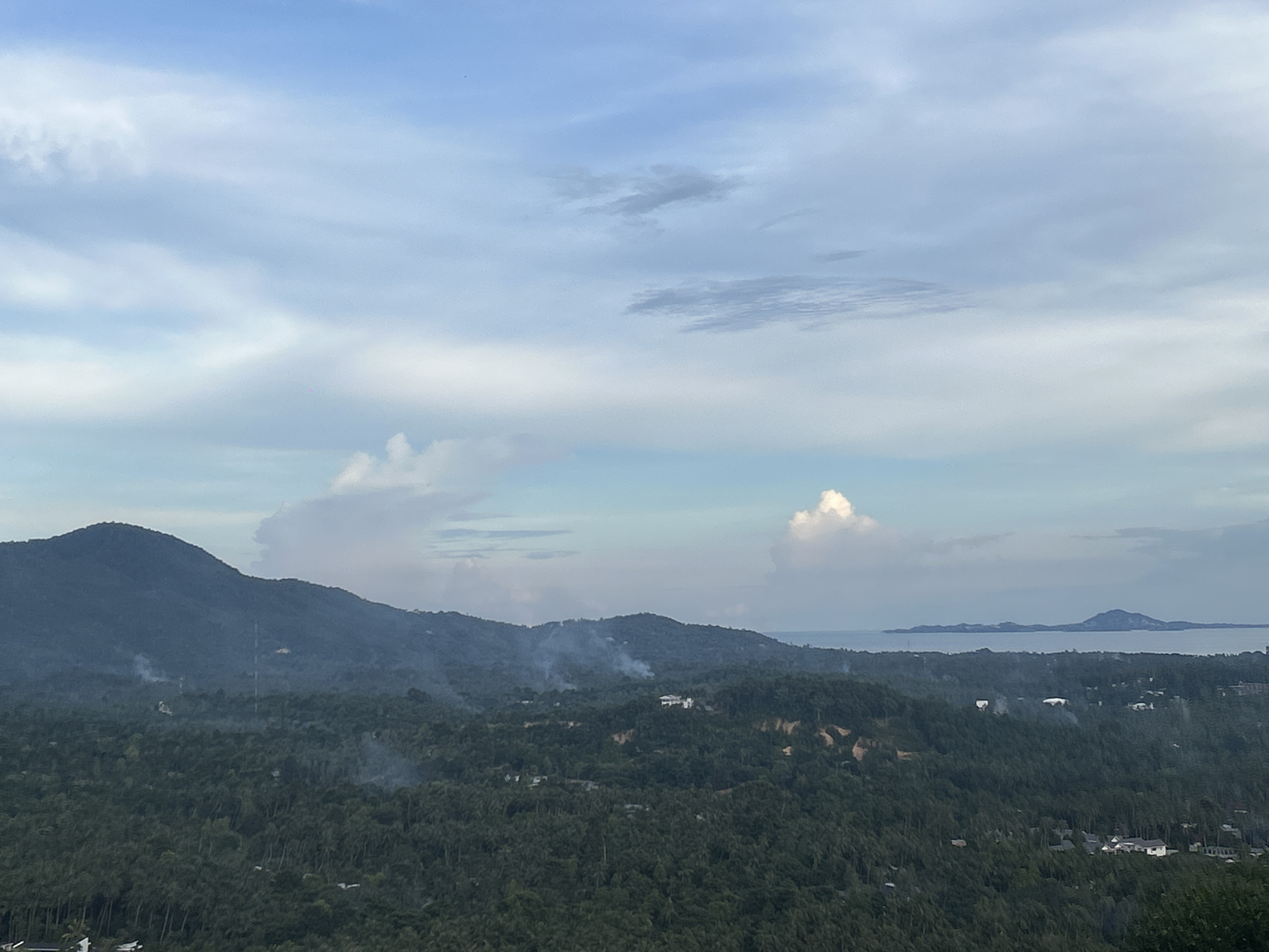 The image shows a landscape scene in Baan Hin Kong, Thailand, featuring a view from an elevated position. The foreground is dominated by dense, dark green vegetation, likely a forest or jungle. Smoke plumes rise in several locations amidst the trees, suggesting possible fires or controlled burns. A mountain range is visible in the midground, extending across the frame. In the background, there is a body of water, with a distant island. The sky is partly cloudy, with a mix of light blue and white clouds.