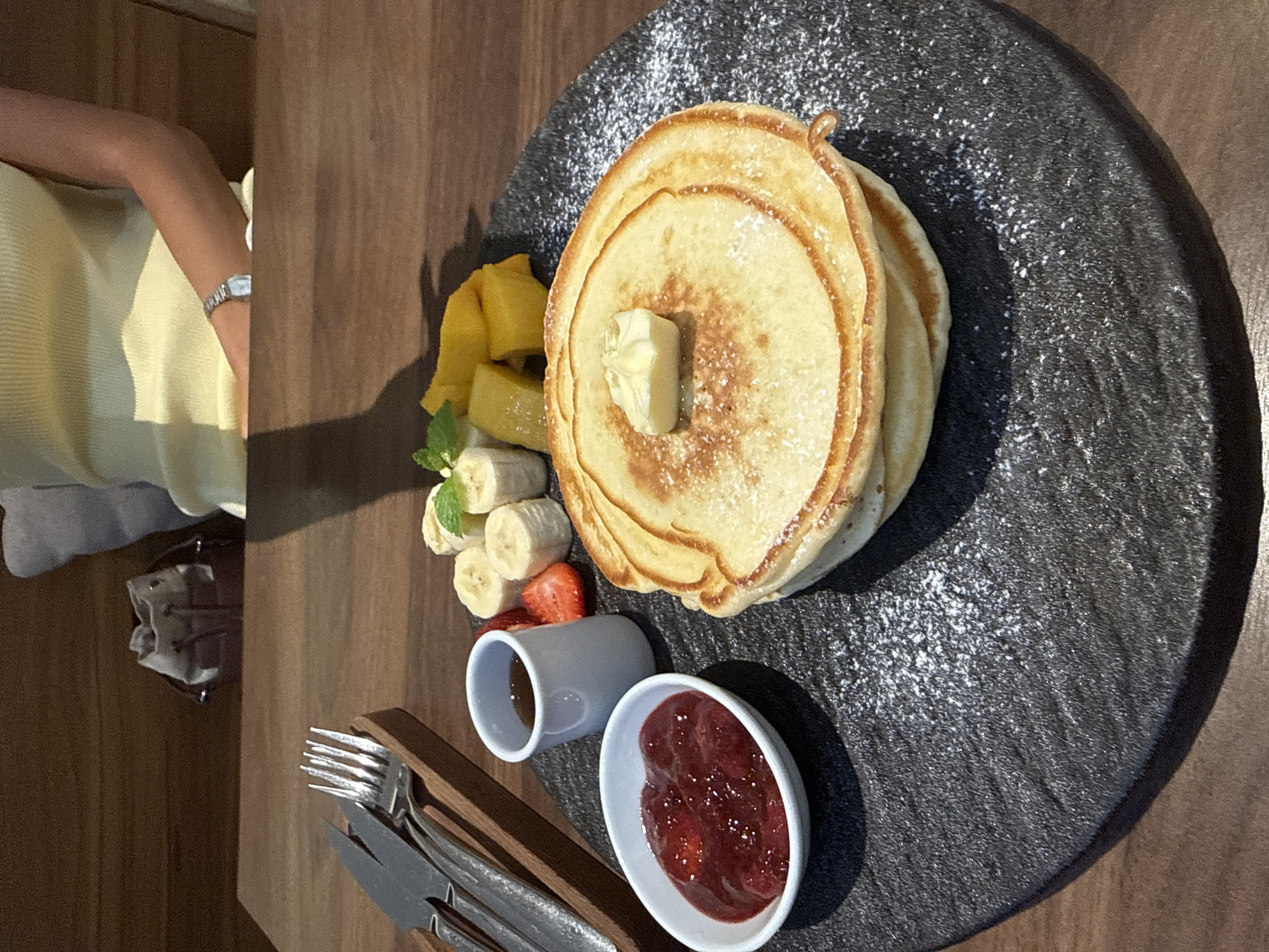 The image presents a top-down view of a plate of pancakes served with fruit and condiments. The pancakes are stacked, topped with a pat of butter and sprinkled with powdered sugar. Sliced bananas, diced mango, and sliced strawberries accompany the pancakes. There are two small white bowls; one appears to contain a jam-like substance and the other a liquid, possibly syrup. A set of silverware rests beside the plate. A person's arm, wearing a watch, is visible in the periphery of the frame. The food sits on a round, textured black plate atop a wooden table. The setting suggests a breakfast or brunch meal at a restaurant. Without additional information, a specific location in Đà Nẵng, Vietnam, cannot be determined.