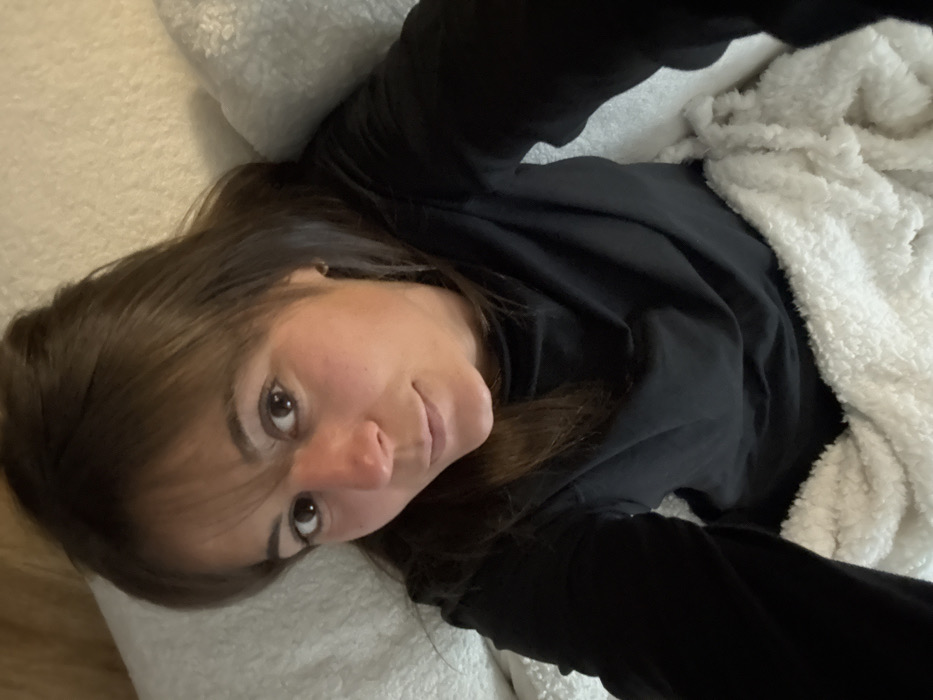 A woman with brown hair and a black shirt is lying on a white, fluffy blanket. She is looking up at the camera with a slight smile. Her head is resting on a white pillow, and her body is partially covered by the blanket. The lighting is soft and natural, and the overall tone of the image is relaxed and cozy.