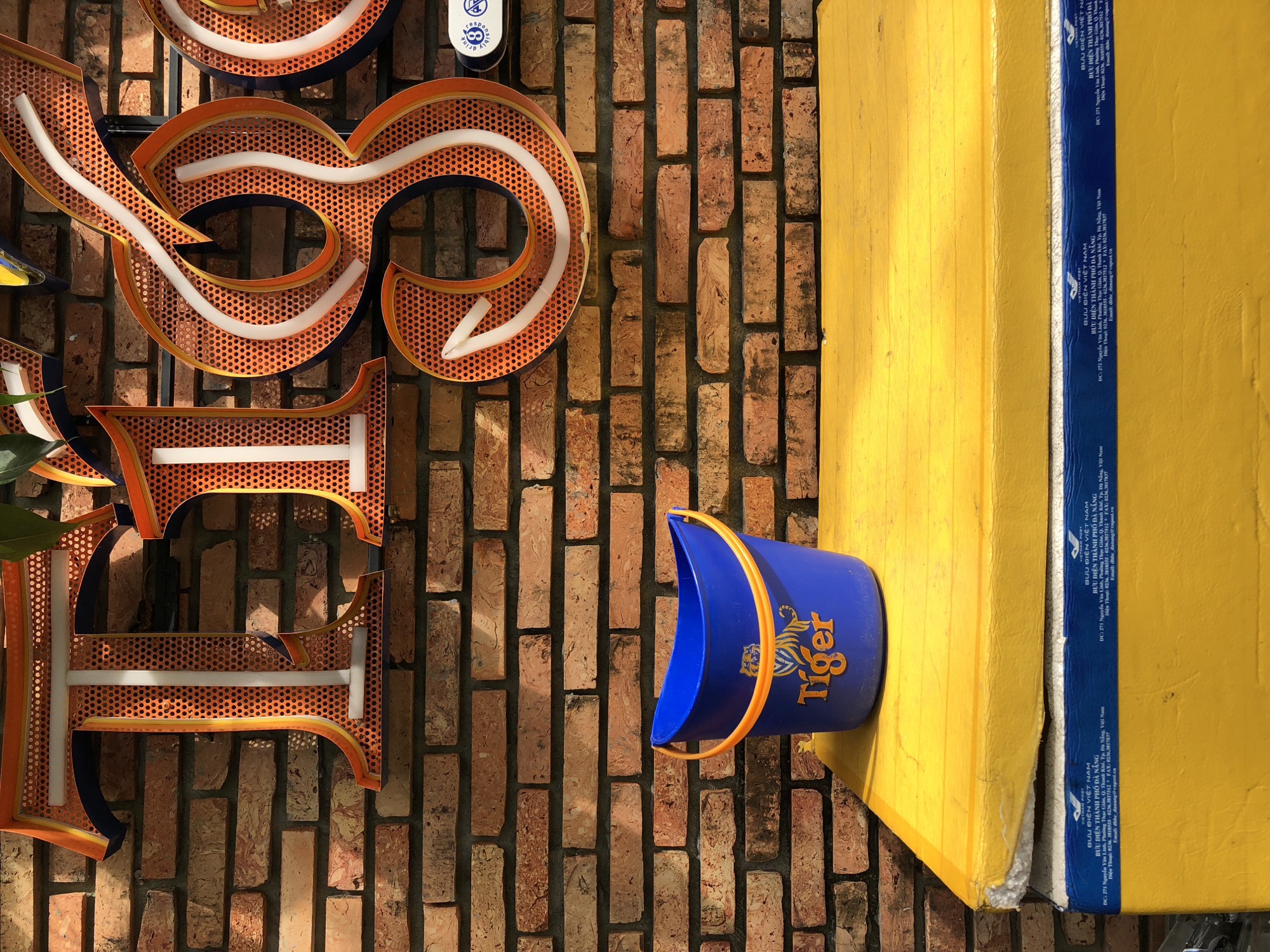 A blue plastic bucket with an orange handle and the "Tiger" logo is placed on a bright yellow surface in front of a brick wall. The wall has unevenly colored and textured bricks. Above the bucket and to the right, there are large, illuminated neon signs on the brick wall. The signs appear to be letters spelling out "Tiger," with orange tubing outlining the letters and white neon strips inside. The bucket has a light reflection on its surface. Below the yellow surface is a cooler with blue and white branding that includes text in Vietnamese, indicating it is from "BƯU ĐIỆN THÀNH PHỐ ĐÀ NẴNG" (Da Nang City Post). The cooler also has text with an address and phone numbers in Vietnamese. The lighting suggests it is daytime, with shadows cast by the bucket and cooler.