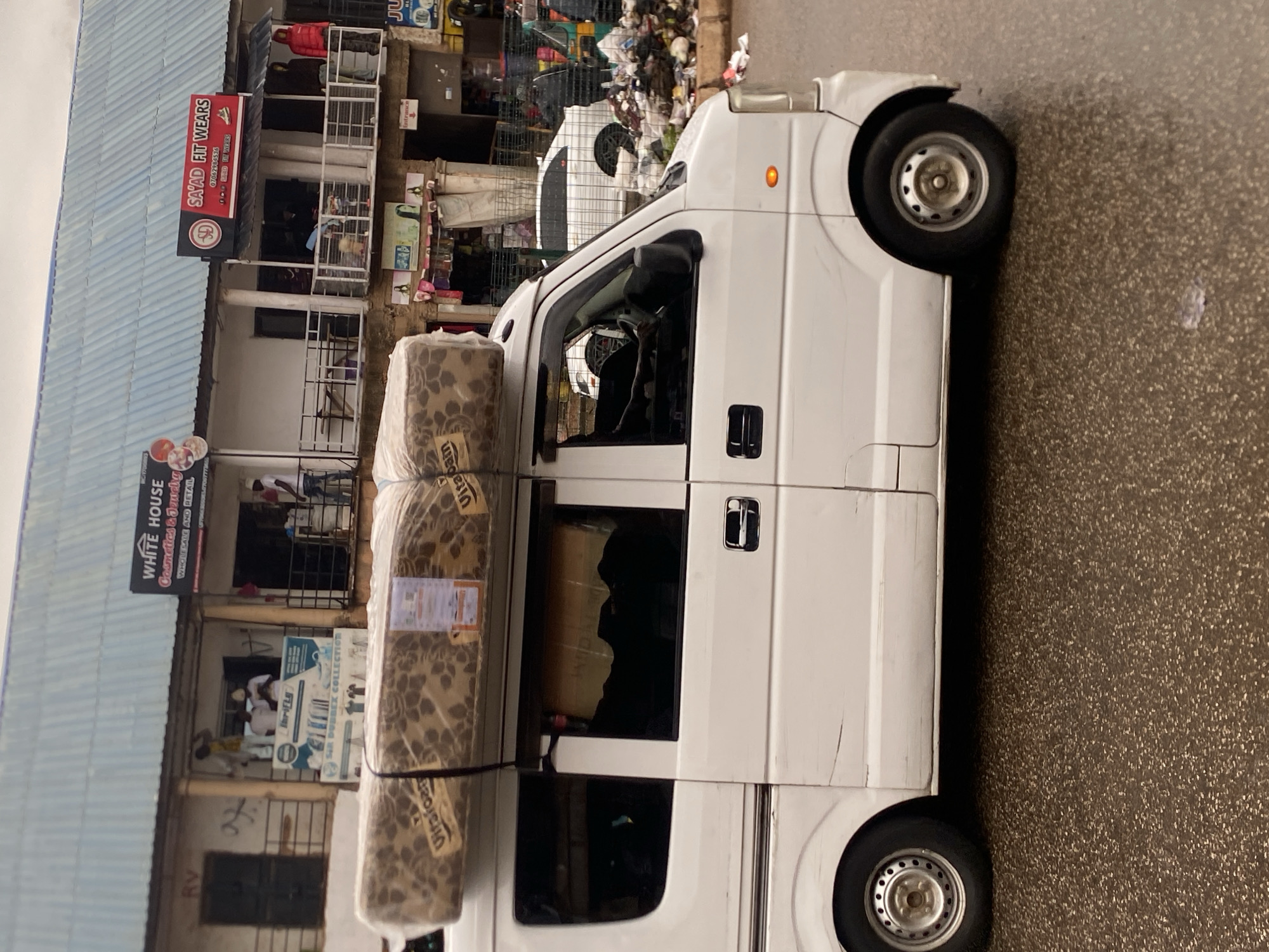 The image shows a white van parked next to a row of shops in Bukuru, Nigeria. The van is loaded with rolled-up mattresses. One of the shops has a sign that reads "SAYAD FIT WEARS" and another reads "WHITE HOUSE Cosmetics & Jewelry". Another sign visible on the van itself reads "Vitafoam". The scene appears to be a commercial street with people visible inside the shops and some items stacked up outside. The weather appears to be overcast. The overall impression is a busy street scene with commercial activity taking place.