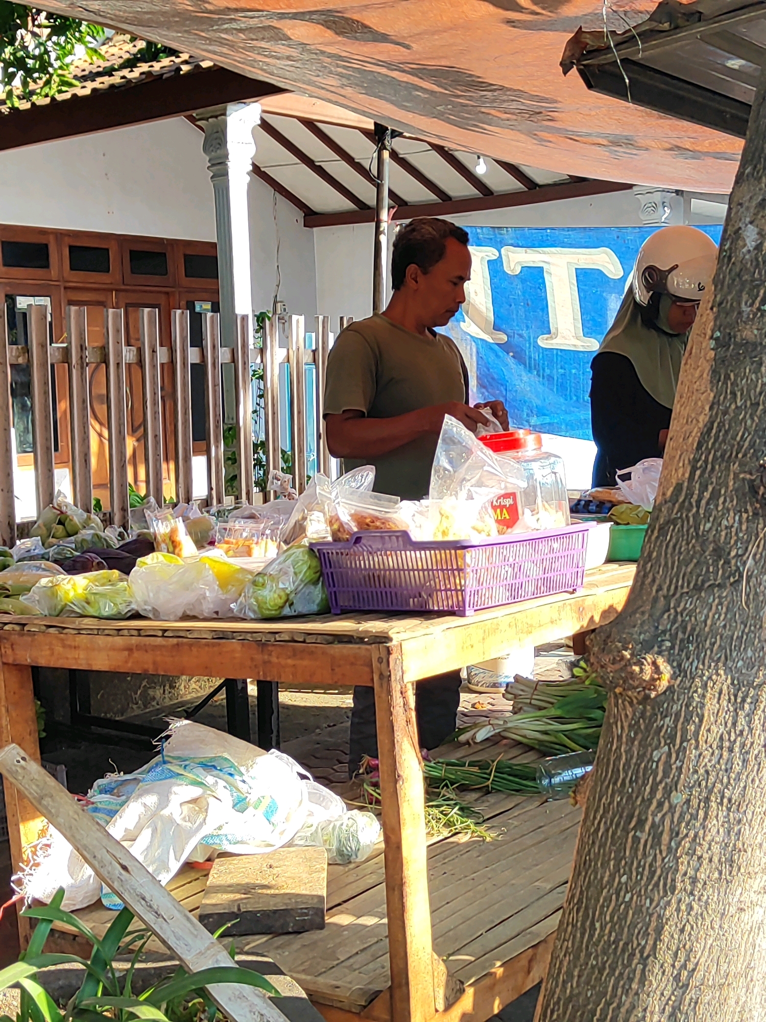 The image captures a street market scene in Bondowoso, Indonesia, under a makeshift canopy. A man in a dark olive t-shirt is standing behind a wooden table laden with produce. He is in the process of handling plastic packaging, possibly for sale. To his right, a person wearing a light green hijab and a white helmet is partially visible, also engaged in market activities. The table displays a variety of items, including what appear to be corn, bananas, and leafy green vegetables, many of which are bagged in plastic. A purple basket containing food items is also prominent on the table. In the background, a building with wooden doors and columns is visible, and a large blue banner with white lettering hangs above the stall. A large tree trunk occupies the right side of the frame, partially obscuring the scene. The ground beneath the table is made of wooden planks.