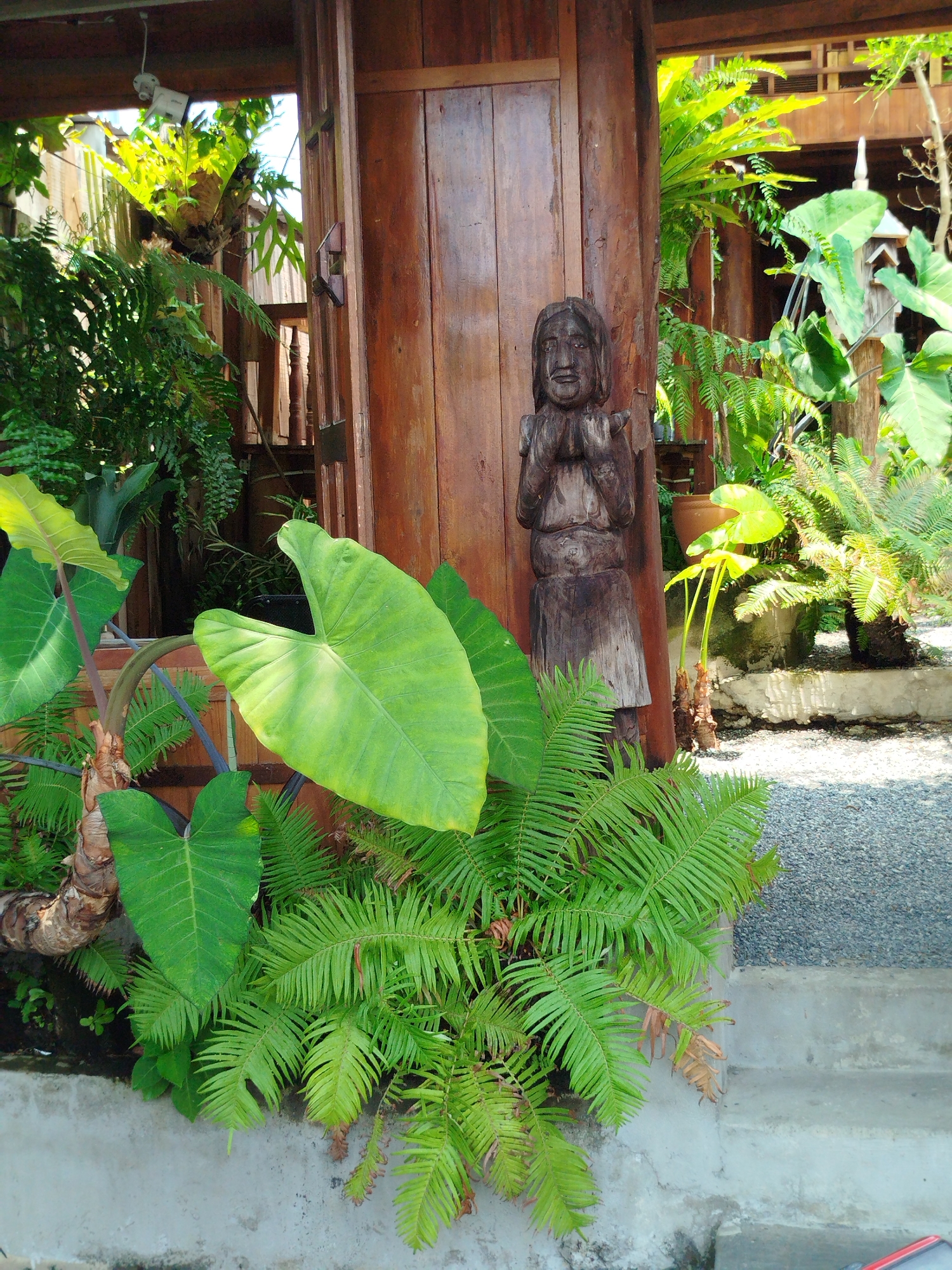 The image depicts a wooden structure in a garden-like setting, possibly in Ho Chi Minh City, Vietnam. A dark wooden carving of a person with hands clasped in prayer stands adjacent to a wooden post. The foreground features lush greenery, including large-leafed plants and ferns. The background suggests more vegetation and wooden architectural elements. The setting appears to be a serene, well-maintained garden.