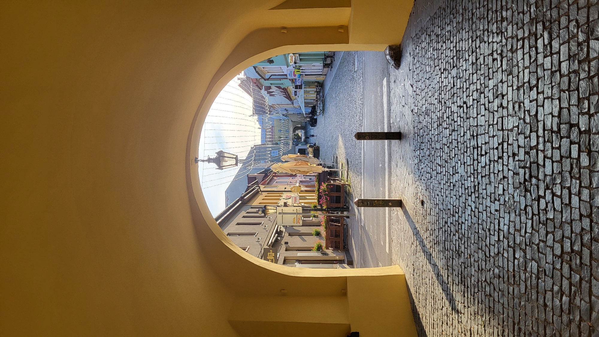 The image captures a view through a yellow archway onto a cobblestone street in Prešov, Slovakia. The street is lined with colorful buildings and appears to be a commercial area with outdoor seating for cafes and restaurants. String lights are strung across the street, suggesting a festive atmosphere or preparation for an event. Two dark bollards stand in the foreground on the cobblestone path. Sunlight casts long shadows across the street, indicating it is either early morning or late afternoon. A single street lamp hangs from the archway. No people are prominently featured, and the scene is generally quiet.