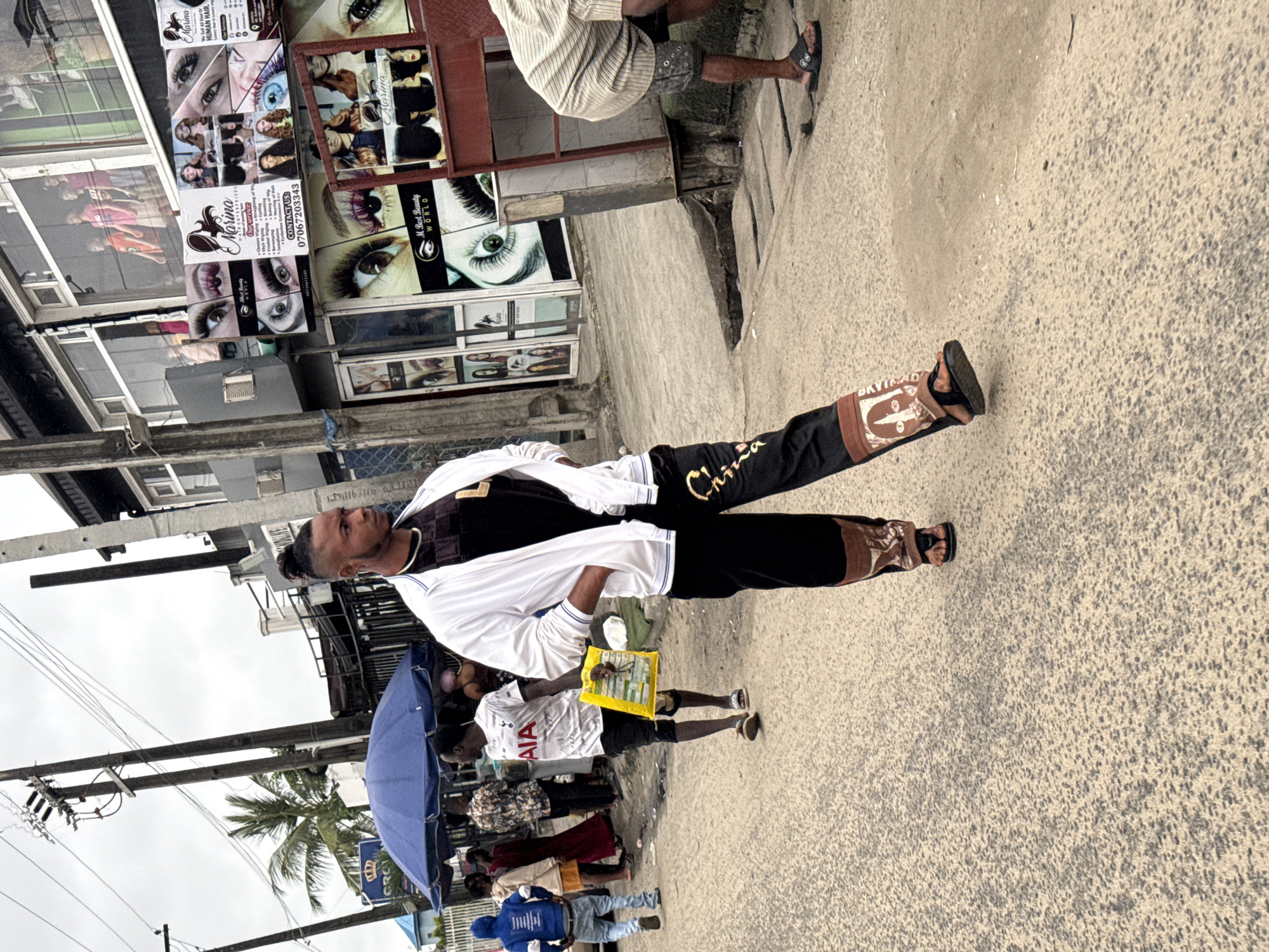 In Ado, Nigeria, a young Black man with a high bun hairstyle walks down a street. He is wearing a white jacket over a black t-shirt, and black pants with a large, decorative design on the lower leg, featuring a face and the word "DAVIDSON". He is also wearing brown sandals. In the background, several other people are visible, including a man in a white t-shirt with "AIA" written on it, carrying a yellow bag, and another person in a blue hooded jacket. To the left of the frame, a blue umbrella shades a market stall. The street is paved with asphalt and shows signs of wear. Buildings with advertisements, primarily for beauty services, line the street. Utility poles and wires are prominent in the overhead view, suggesting an urban environment. The sky is overcast.