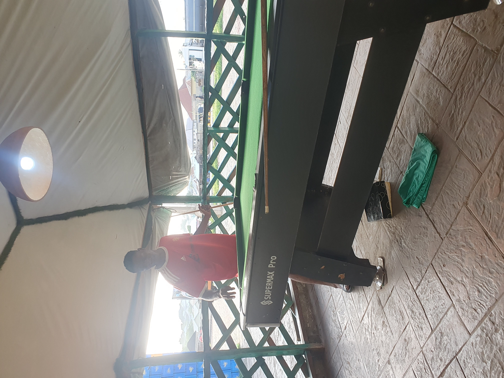 An overhead shot captures a young man standing next to a billiard table within a covered outdoor area. The man, wearing a red t-shirt, faces the camera with his left hand resting on the billiard table. The table itself is dark and features the "SUPERMAX Pro" logo. A pool cue lies on the table. The ground is paved with stone tiles. In the background, a wooden fence separates the billiard area from an open space with buildings and vehicles. The scene appears to be a recreational area, possibly a bar or restaurant, located in Lokoja, Nigeria.