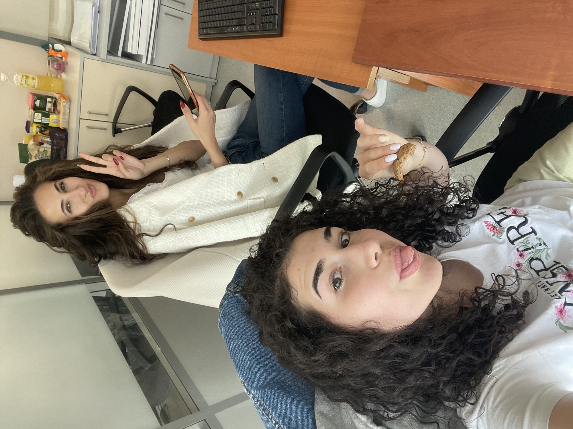 Two young women pose for a selfie indoors, likely in an office environment. The woman in the foreground wears a white t-shirt with floral lettering and curly dark hair, while the other has dark, wavy hair. The scene includes office furniture: desks, chairs, and a keyboard are partially visible. Storage shelves and containers of food are present in the background. The location is identified as Sofia, Bulgaria.