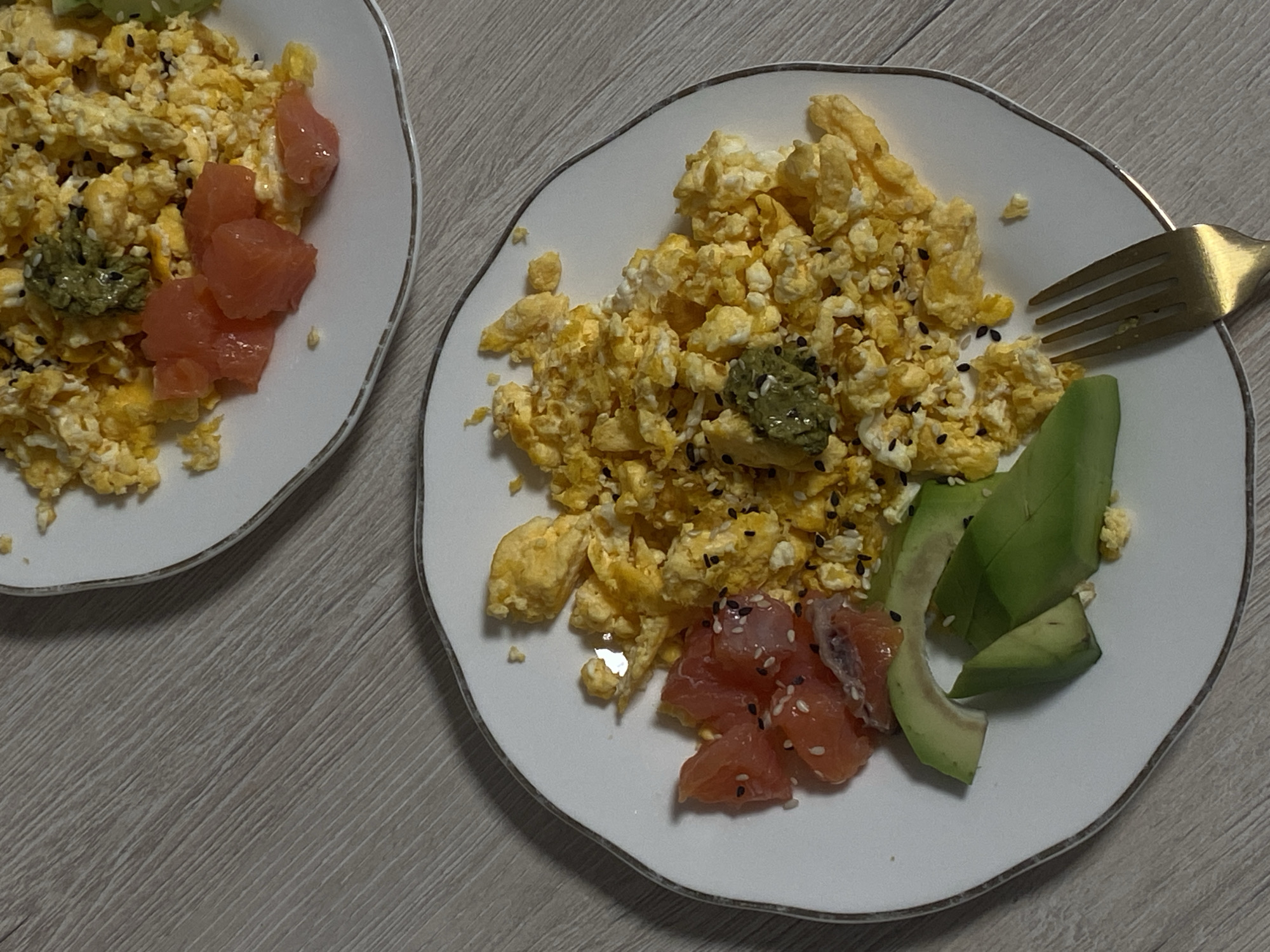 The image shows two plates of food on a light wooden surface, likely a table. Each plate contains scrambled eggs, a portion of salmon, and a dollop of a green paste, possibly wasabi or avocado. One plate also has sliced avocado. Black sesame seeds are sprinkled on the eggs and salmon. A gold-colored fork rests on the plate with avocado. The image does not show any clear indicators that it was taken in Vysokoye, Russia, or any other specific location. There are no people visible in the image.