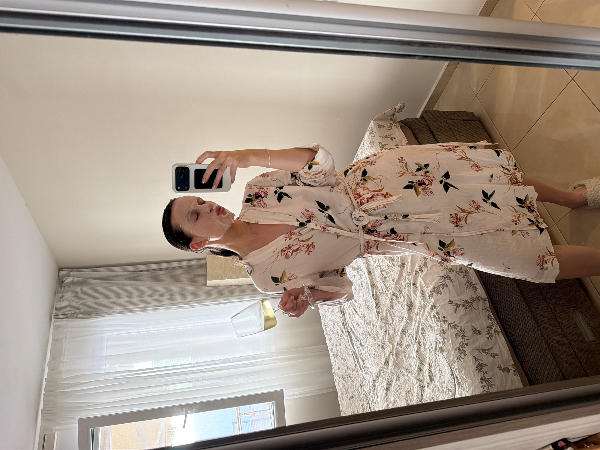 In Budva, Montenegro, a woman with a white sheet mask on her face and her hair pulled back is taking a selfie in a mirror. She is wearing a light-colored floral robe and holding a glass of white wine in her left hand. Her right hand is holding a smartphone. The background shows a bedroom with a bed covered in a patterned comforter, a window with sheer curtains, and a tall mirror with a silver frame. The lighting suggests it is daytime, with sunlight coming through the window.