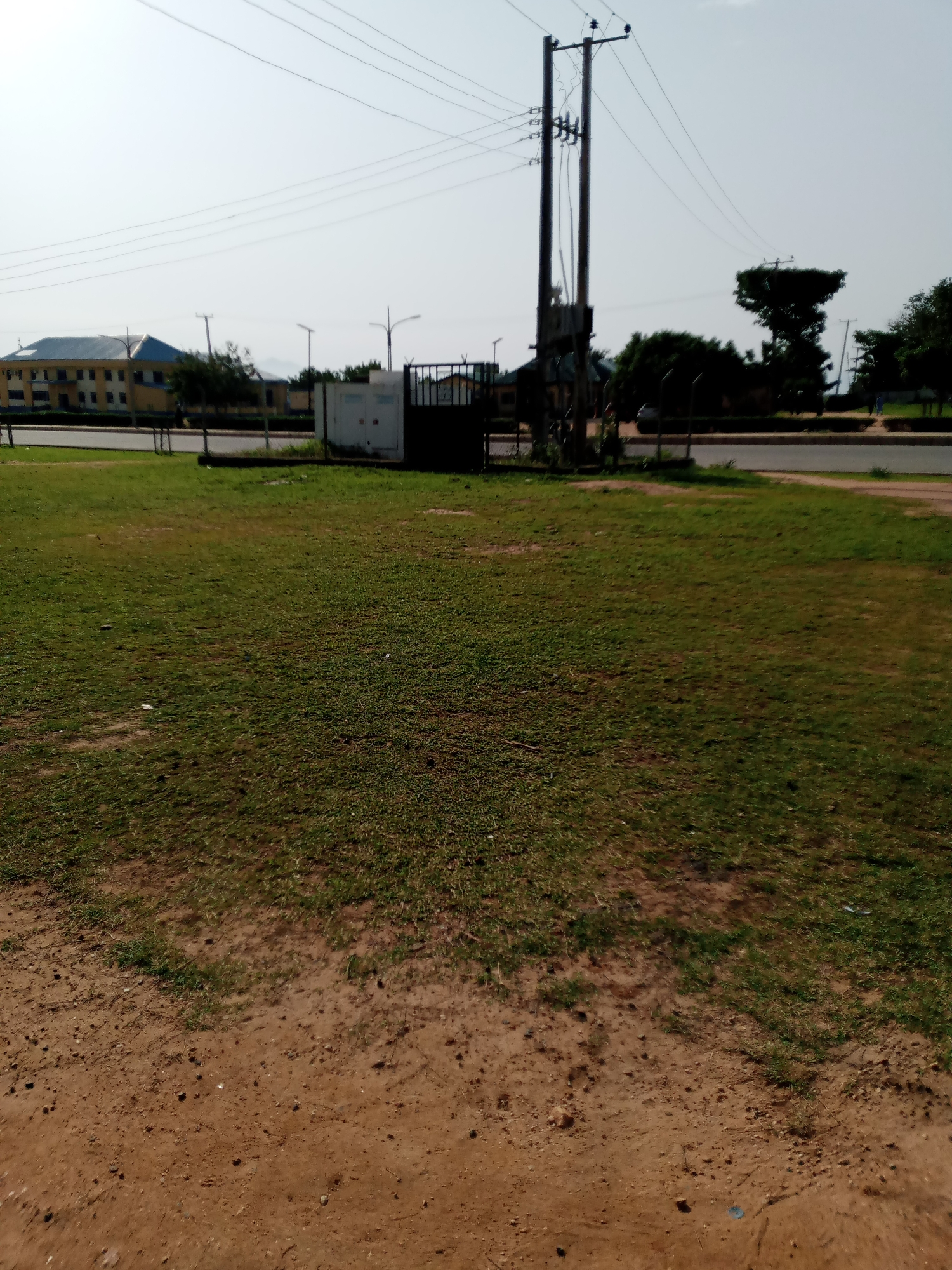 The image shows a grassy field in front of a road with a large, two-story building in the background. A tall electricity pole with wires is visible on the right side of the frame. There is a metal gate and a small white structure next to the electricity pole. The sky is clear and bright. This scene is located in the city of Jalingo, Nigeria.