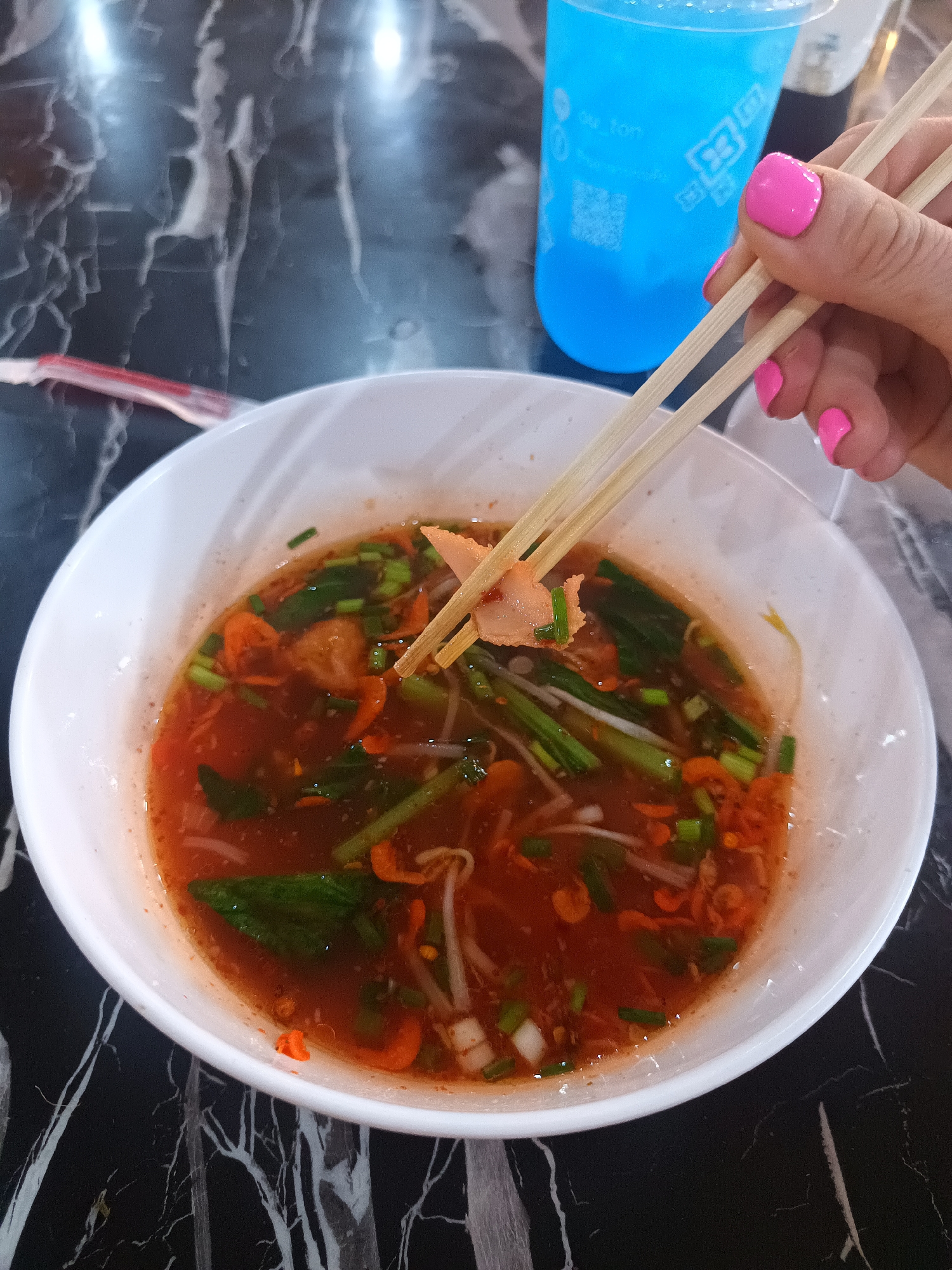 A high-angle close-up shows a bowl of Thai noodle soup held in chopsticks above a black marble-patterned table. The soup consists of a reddish broth with bean sprouts, green vegetables, orange garnishes, and sliced meat. The chopsticks, light brown and made of wood, hold a slice of meat. A hand with pink painted fingernails grips the chopsticks. A blue beverage container is located in the upper right of the frame. A partially visible straw wrapper is present on the left. The scene appears to be set at a restaurant in Thap Sakae, Thailand.